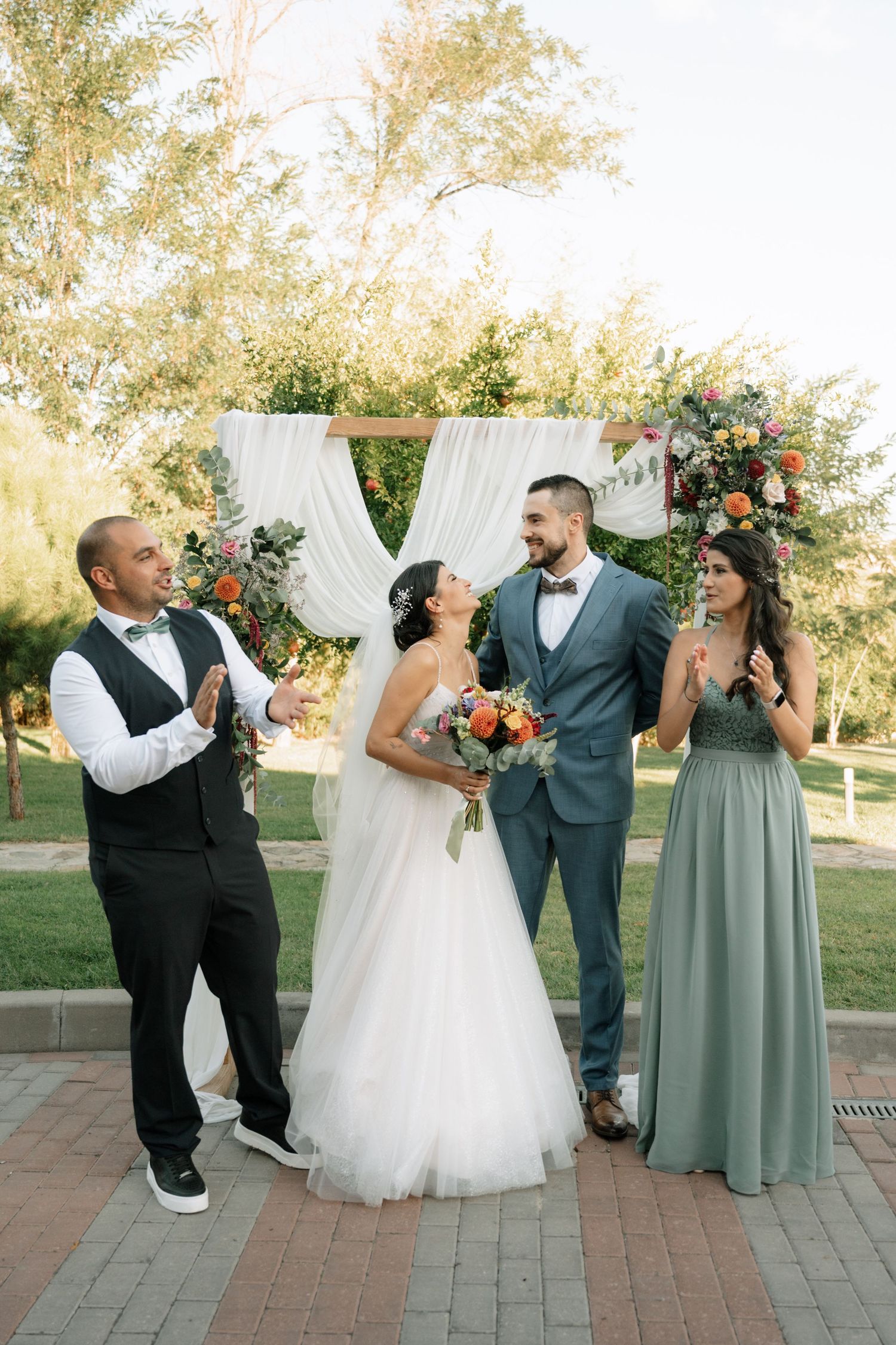 Wedding party poses together on brick patio with white curtains and greenery in outdoor garden setting.