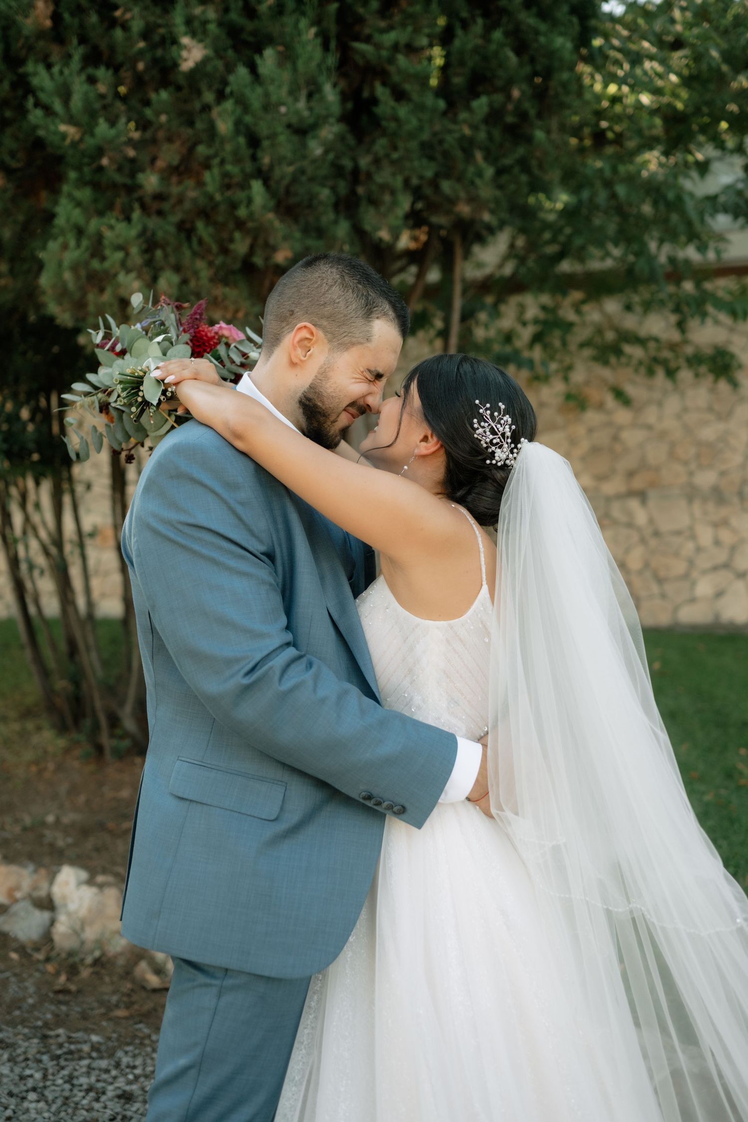 A romantic wedding couple embraces outdoors with the bride in white and groom in blue suit against a garden backdrop.