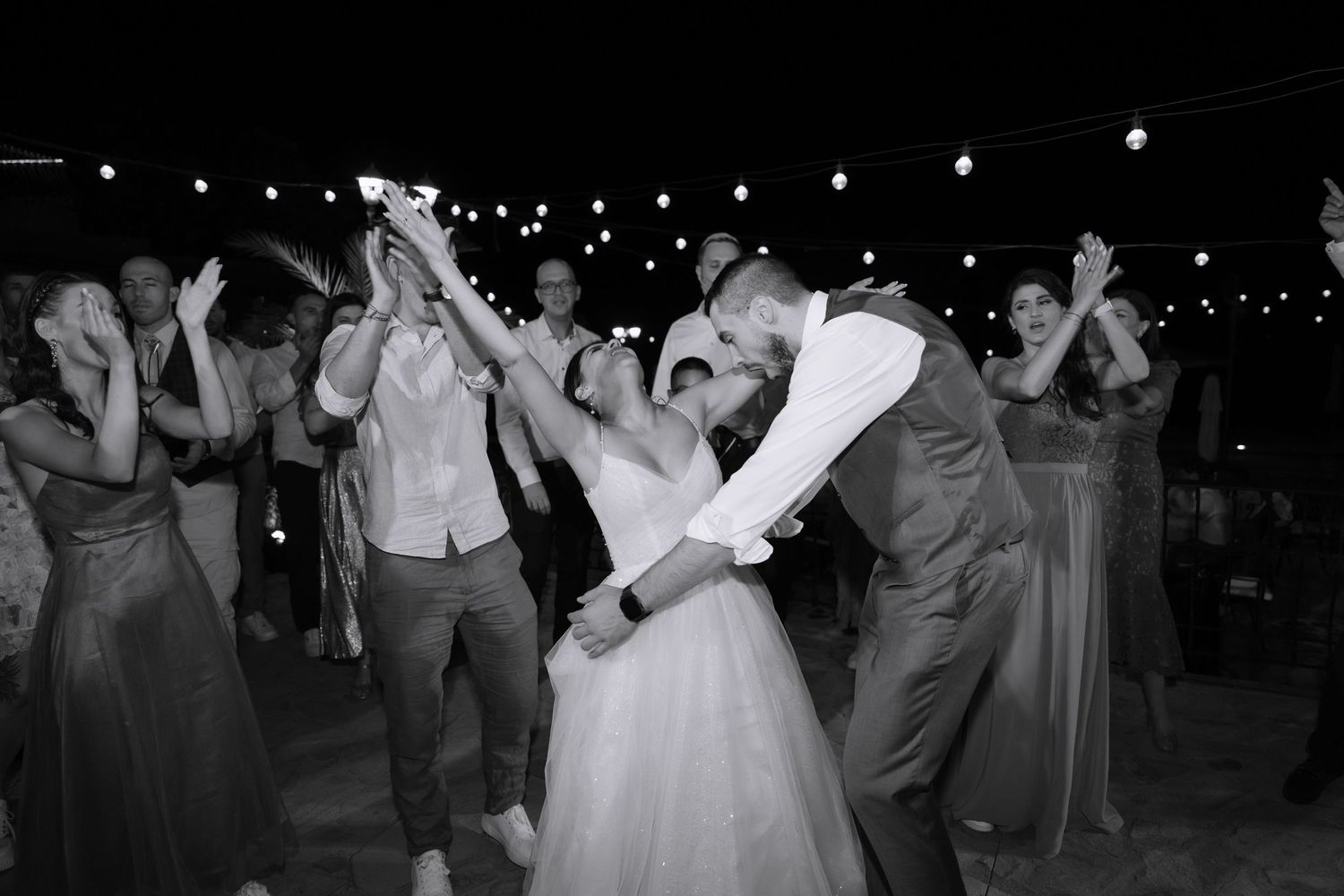 Wedding party dances outdoors at night under string lights during a festive reception celebration.