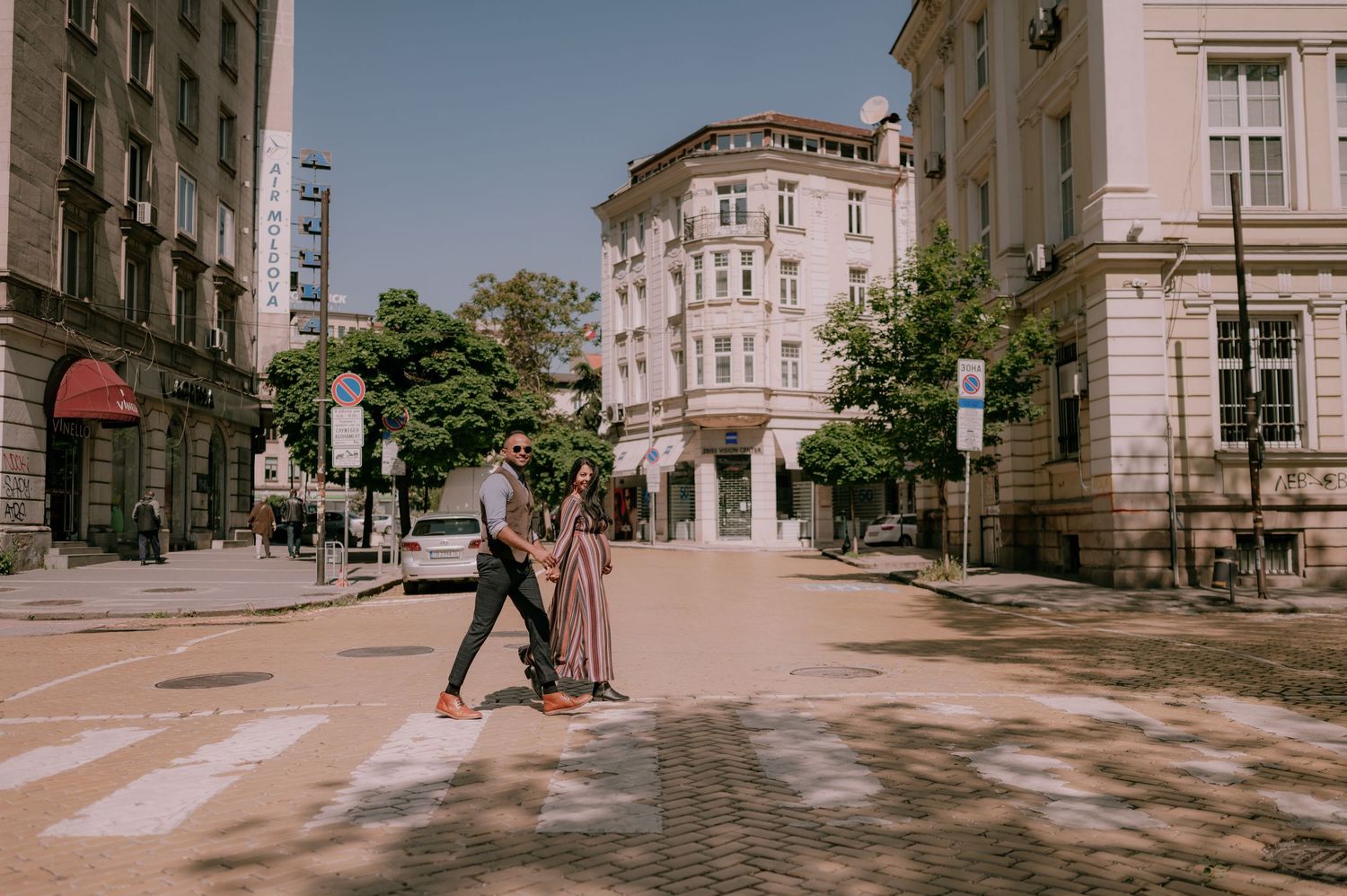 Couple crossing a sunny urban street on a zebra crossing, surrounded by European-style buildings and trees.
