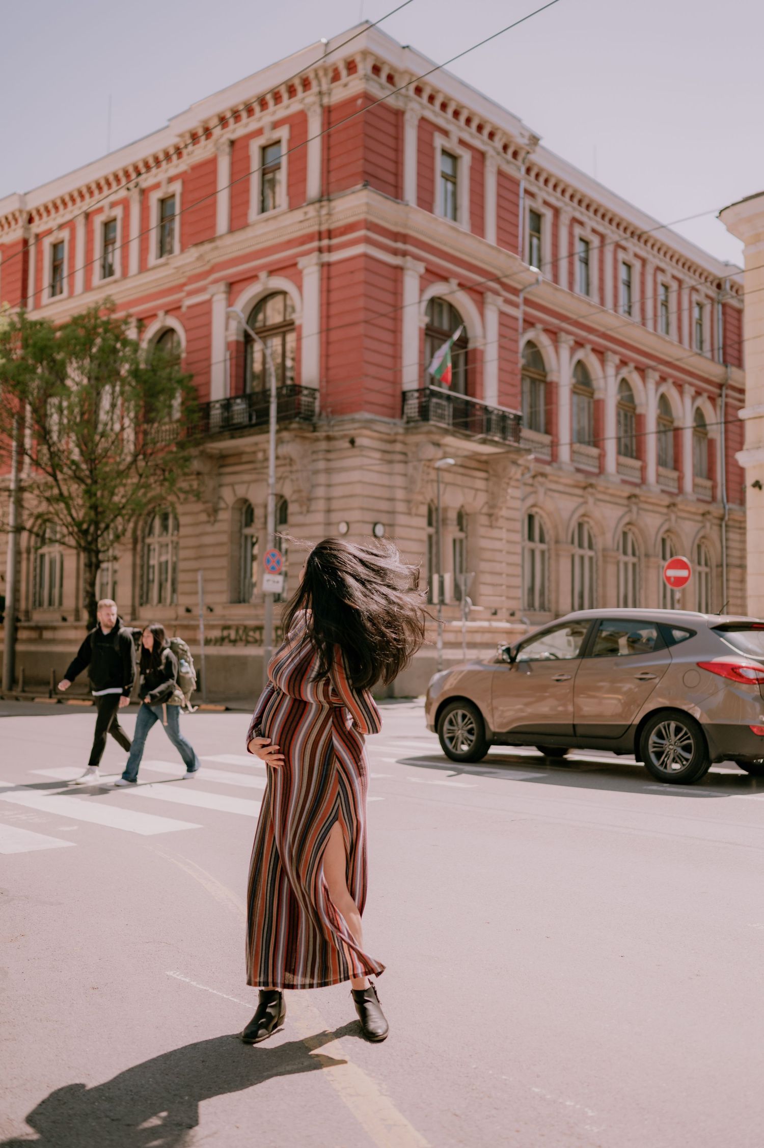 Woman in striped maxi dress crossing urban street with red historic building and passing car in background.