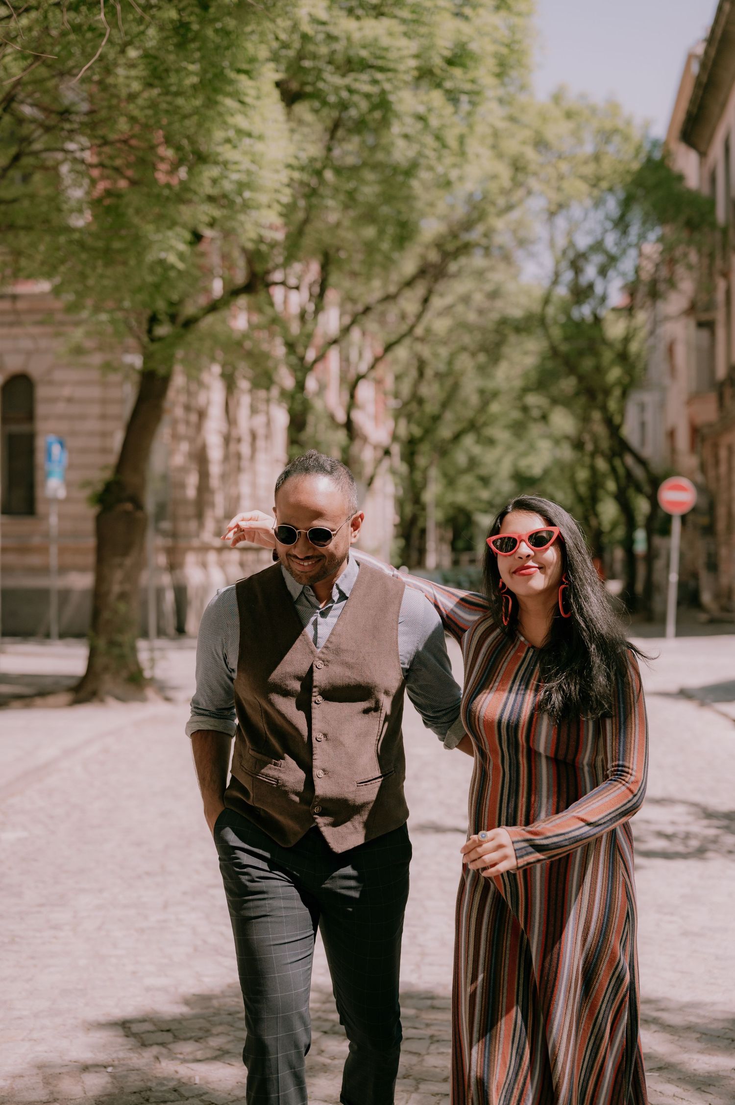 Stylish couple walking outdoors in sunglasses, woman in striped dress, man in vest, on a sunny tree-lined street.