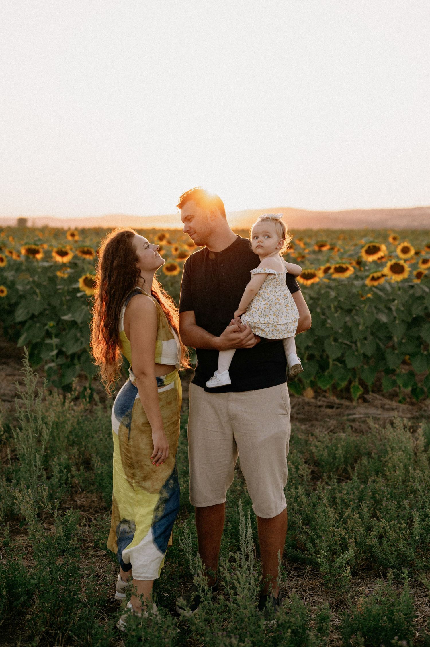 Family of three posing in a sunflower field at golden hour, parents smiling at baby in father's arms.