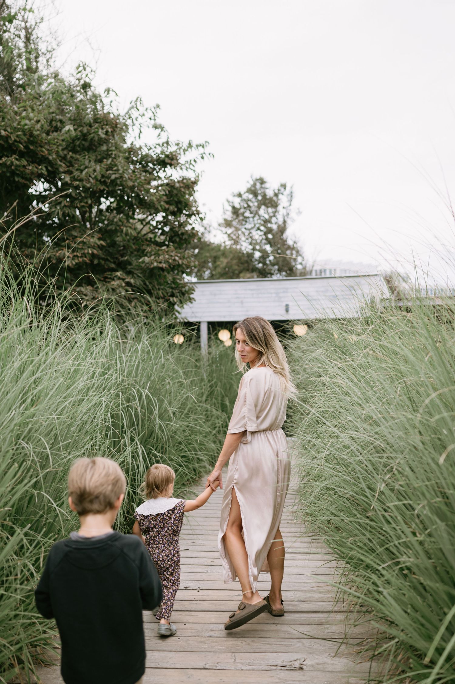 A woman in a beige dress holds hands with two young children on a wooden boardwalk surrounded by tall green grasses.