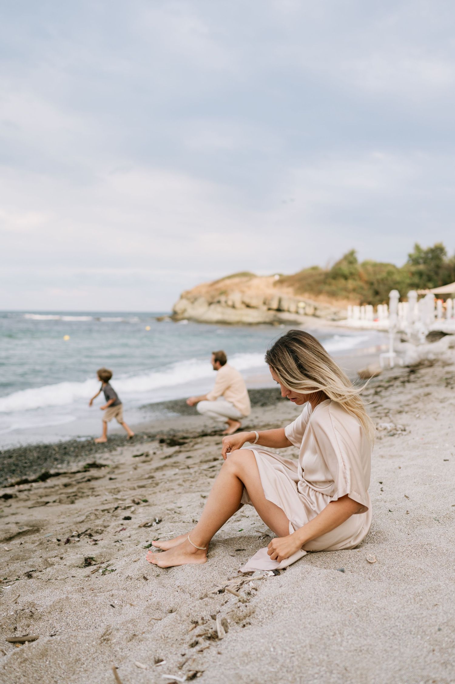 Woman in beige dress sitting on sandy beach while two people play near ocean waves with rocky headland behind.
