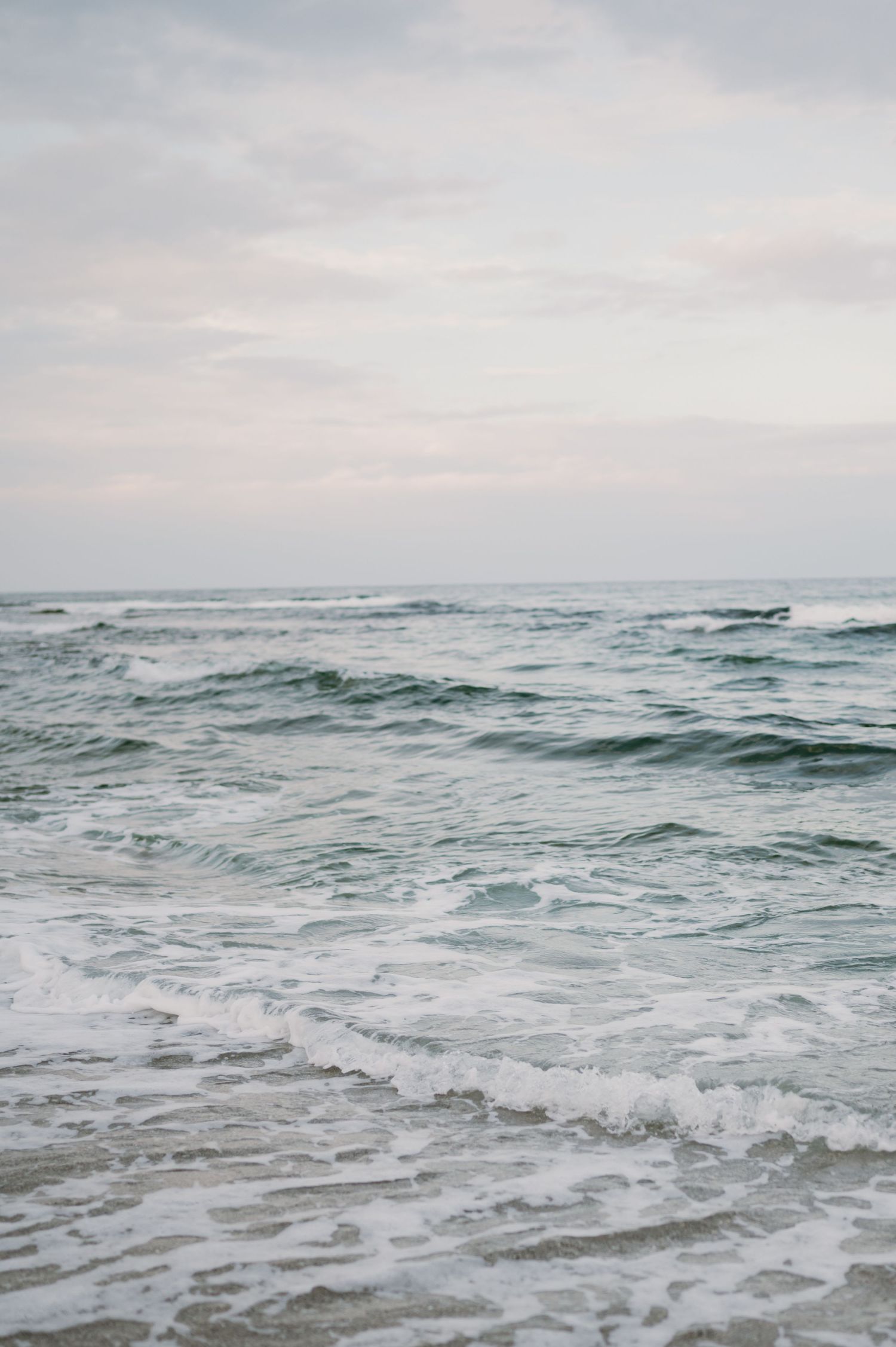 Gentle ocean waves washing onto a sandy beach under a soft cloudy sky, showcasing serene coastal scenery.