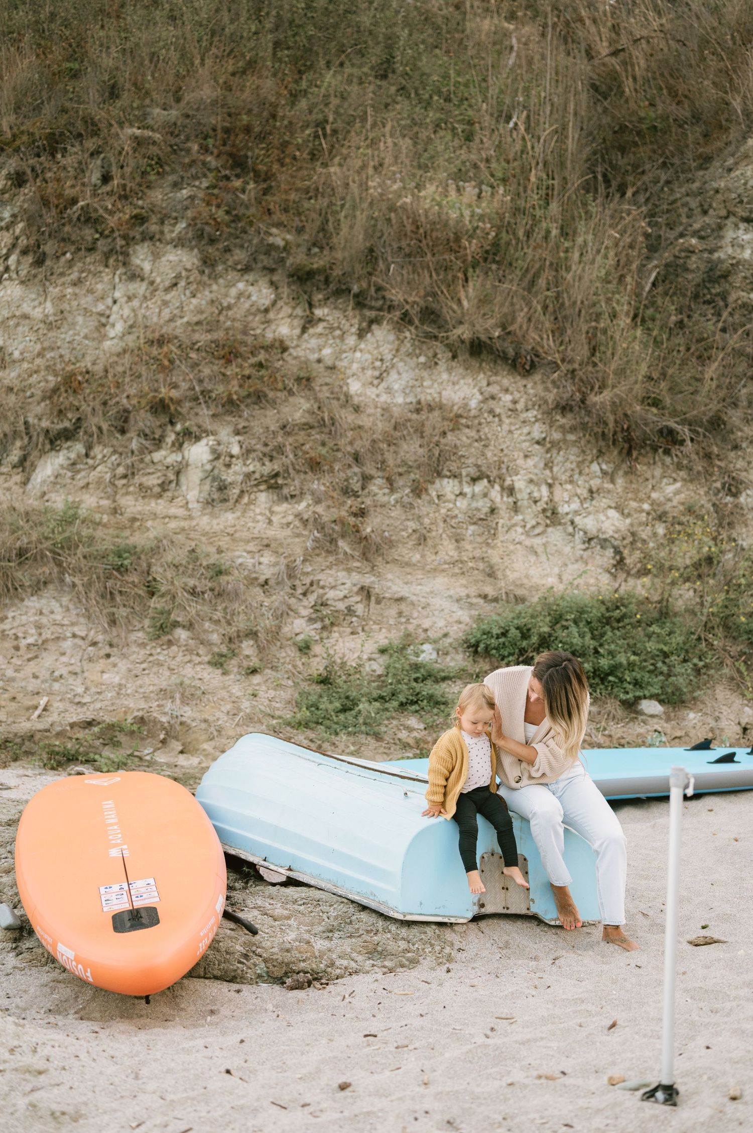 A woman and child sit together on a light blue kayak on a sandy beach near an orange paddleboard and grassy dunes.