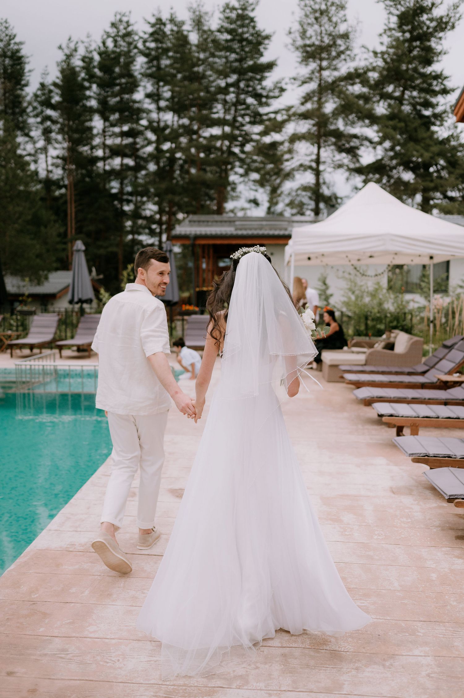 A couple in white wedding attire dances playfully on a pool deck with pine trees in the background.