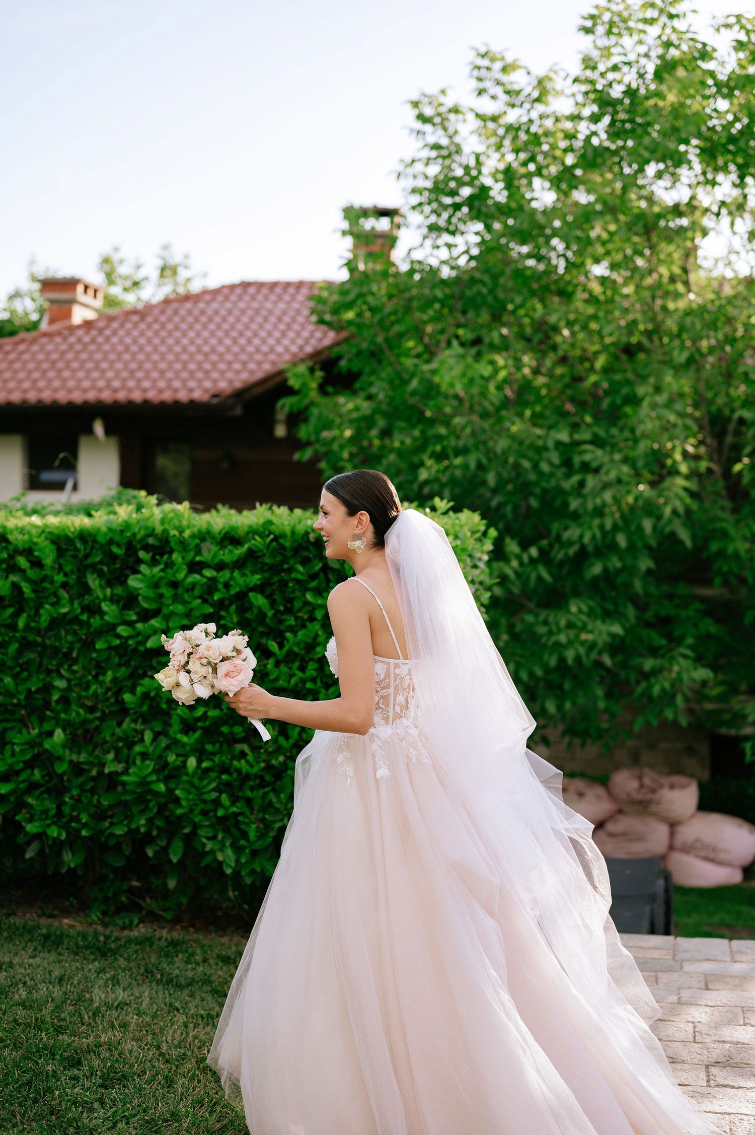 Bride in a lace detailed gown holds bouquet while posing in garden setting.