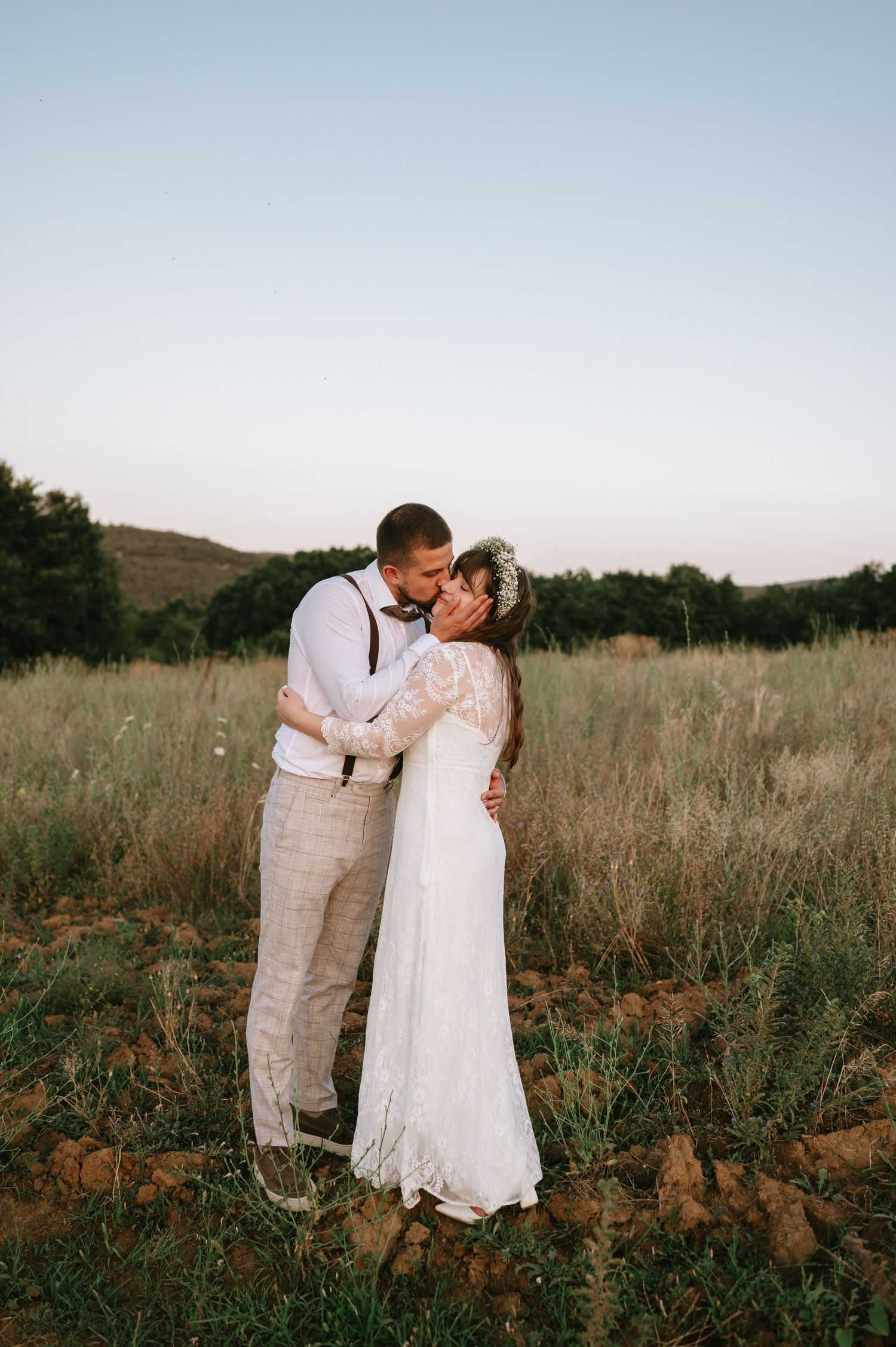 A romantic wedding couple embraces in a rustic field at sunset wearing vintage-inspired attire.