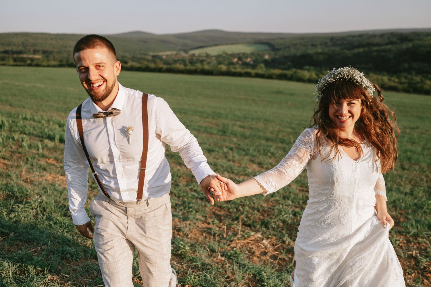 A couple in white wedding attire shares a romantic moment on a hilltop with scenic mountain views.