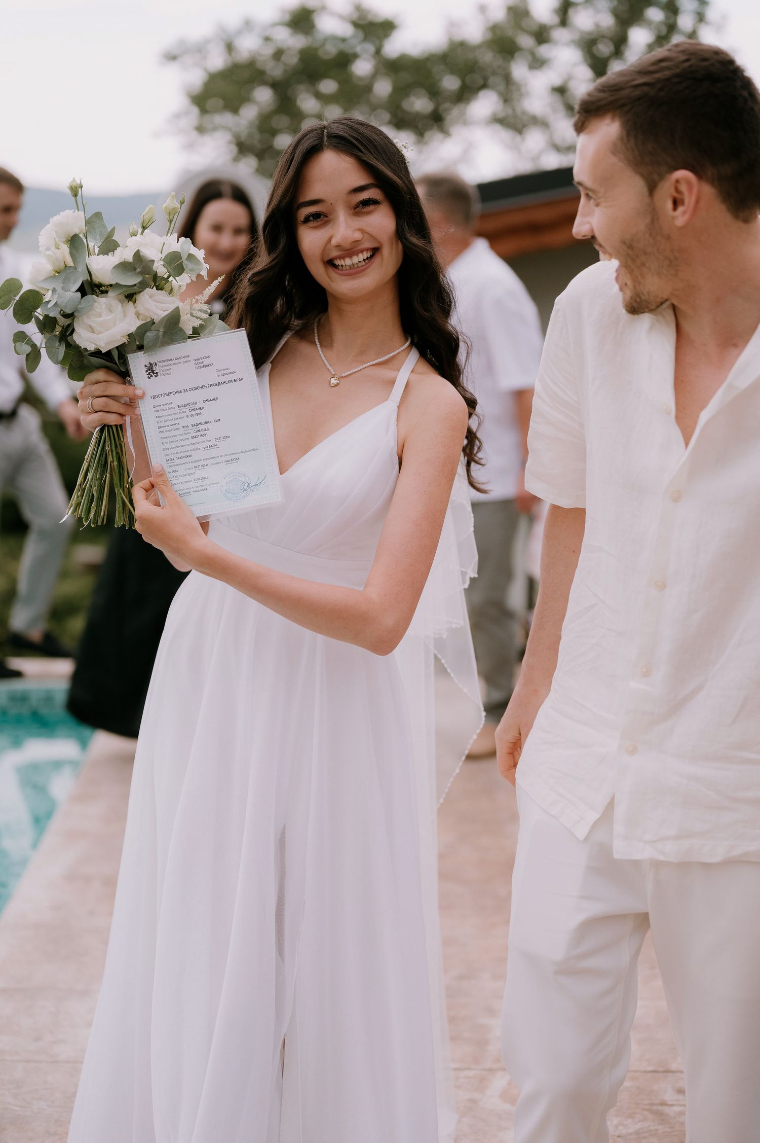 A romantic wedding scene with a couple in all-white outfits holding a bouquet and documents.