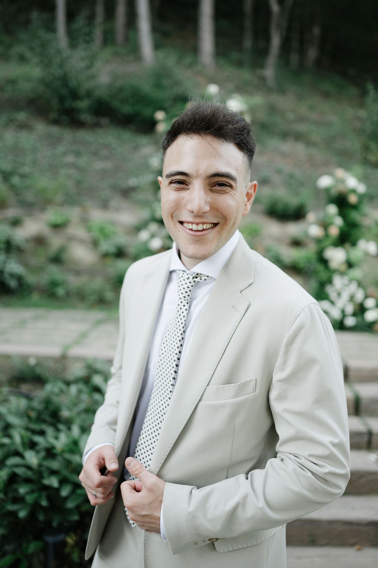 Person in a cream-colored suit smiles against a forest backdrop with white flowers.