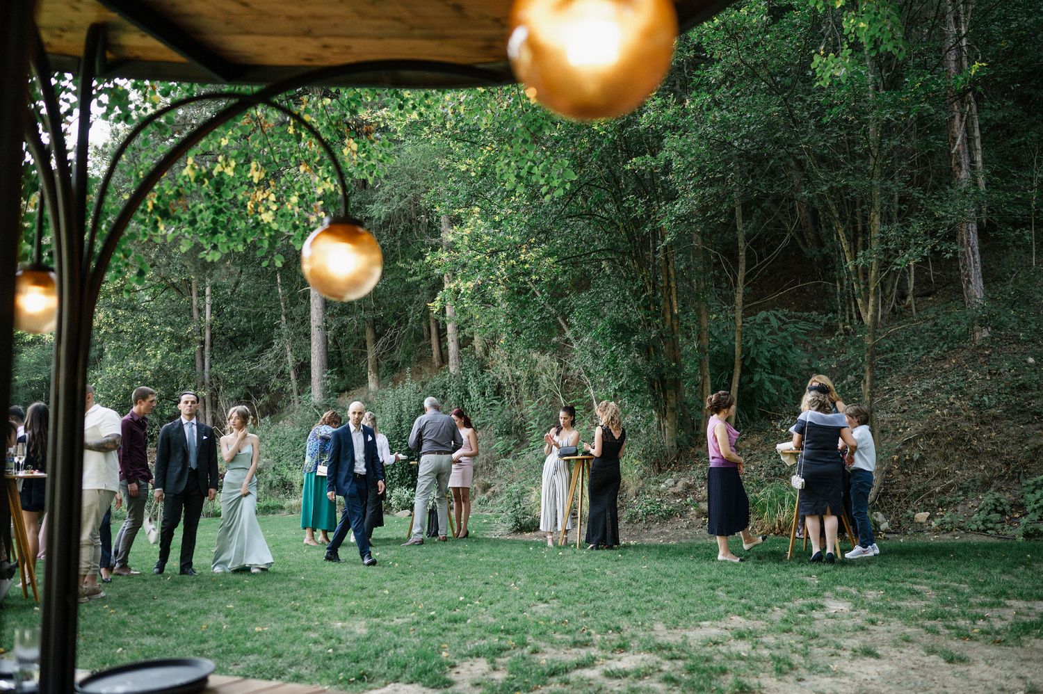 Evening wedding reception in wooded area with hanging golden lights creating magical atmosphere.