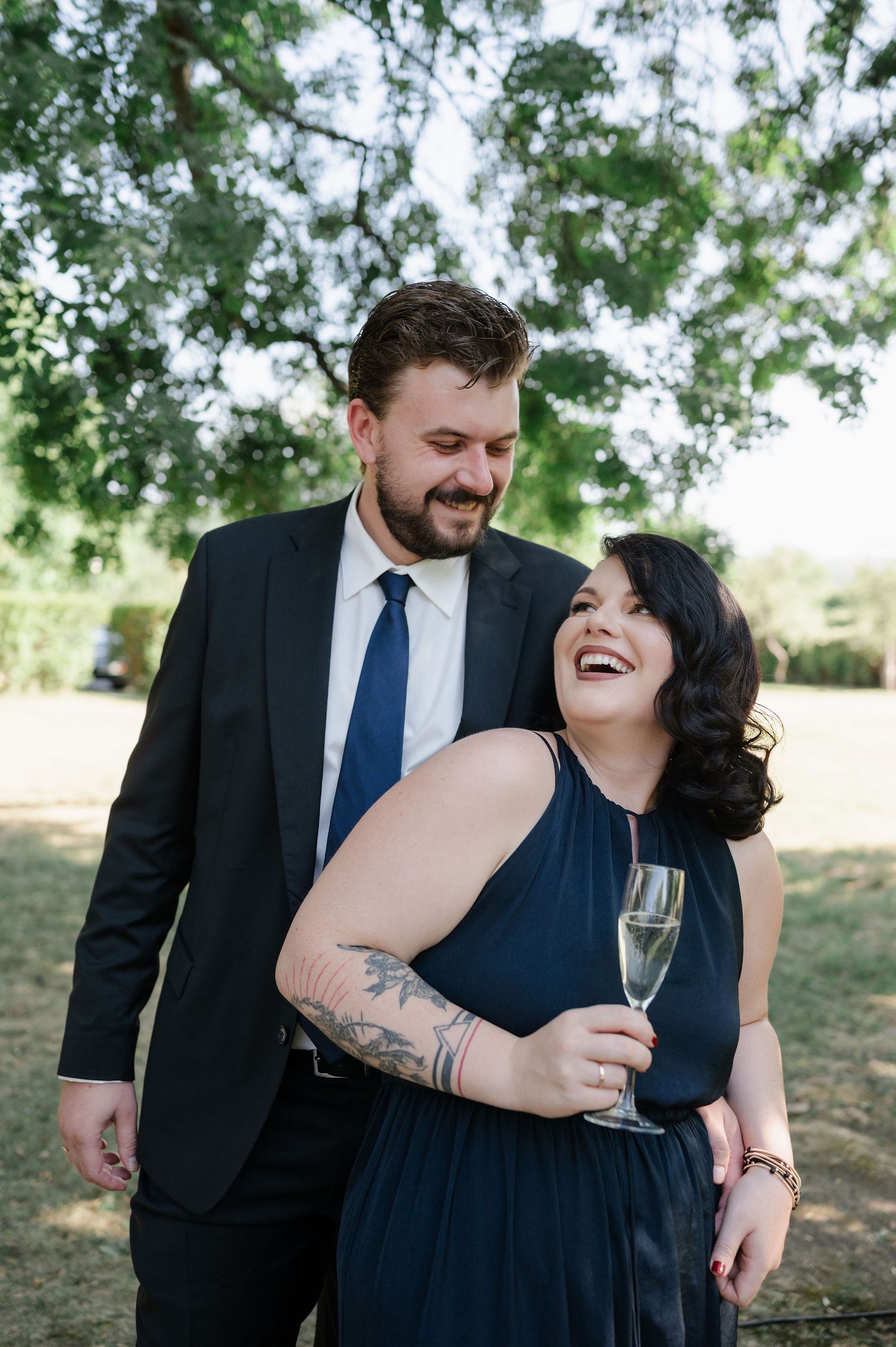 Wedding guests share laughter while holding champagne glasses under a tree canopy.