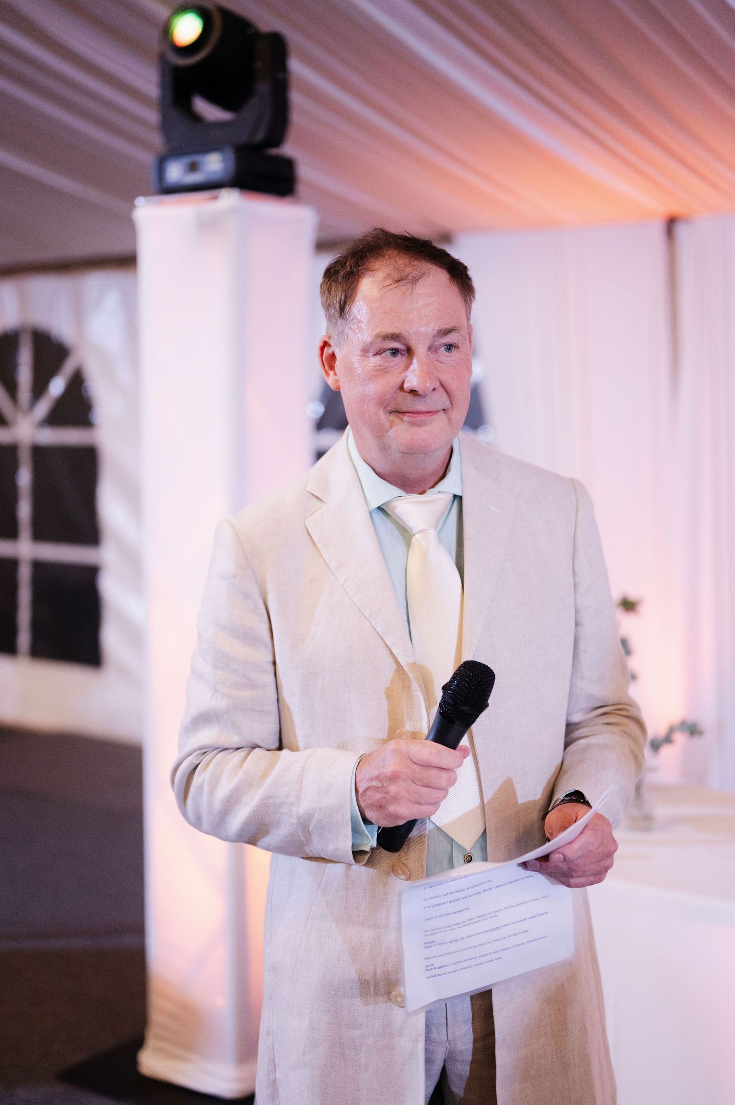 A speaker in a cream-colored suit addresses guests during an evening reception.
