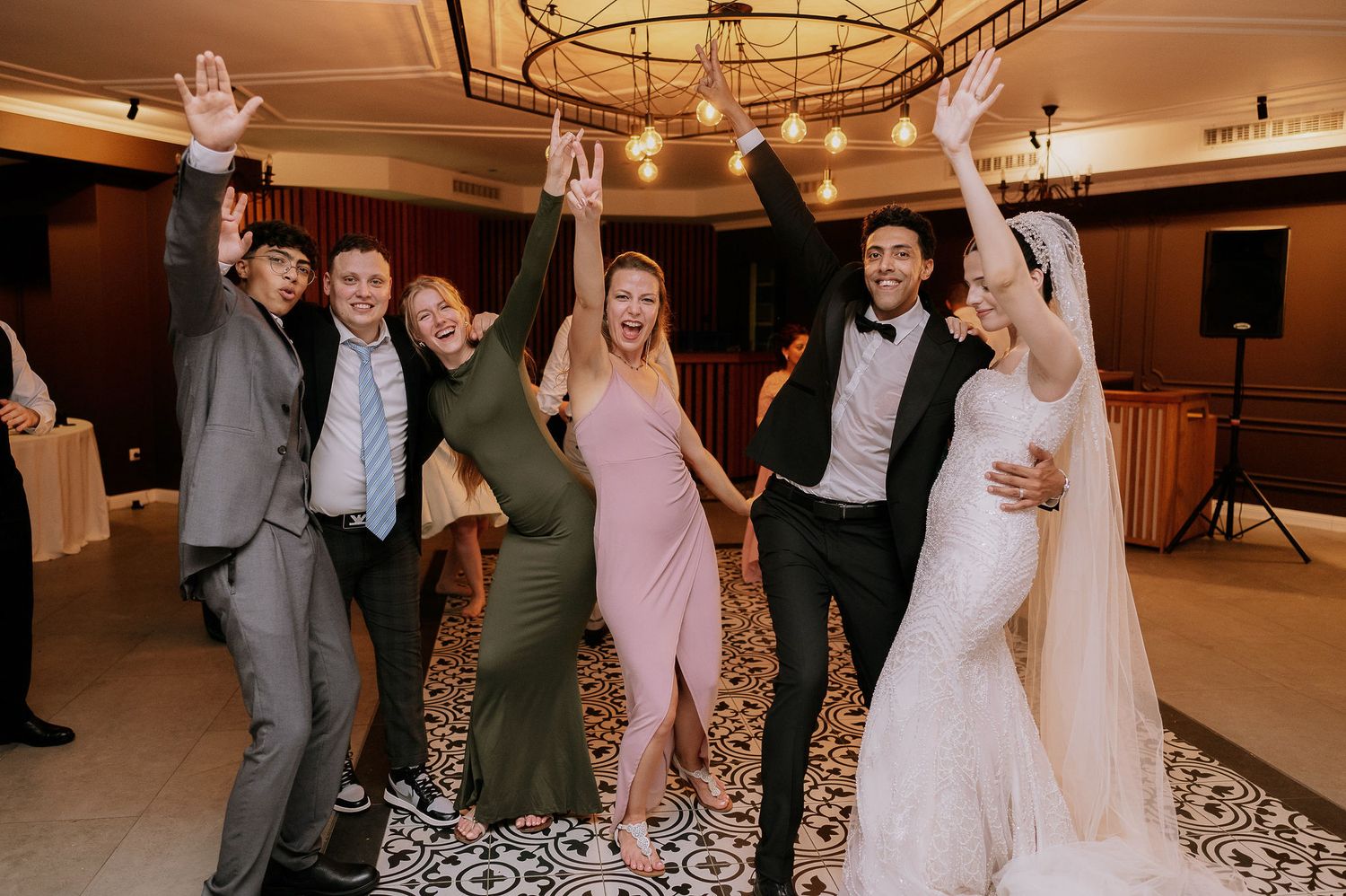 Wedding party poses together in a celebratory moment in a reception venue with ornate lighting.