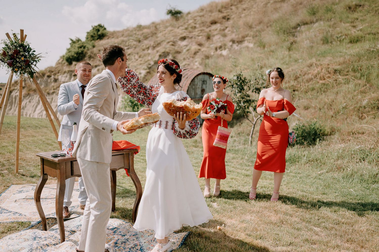 Outdoor wedding ceremony scene with bridesmaids in red dresses and natural hillside setting.