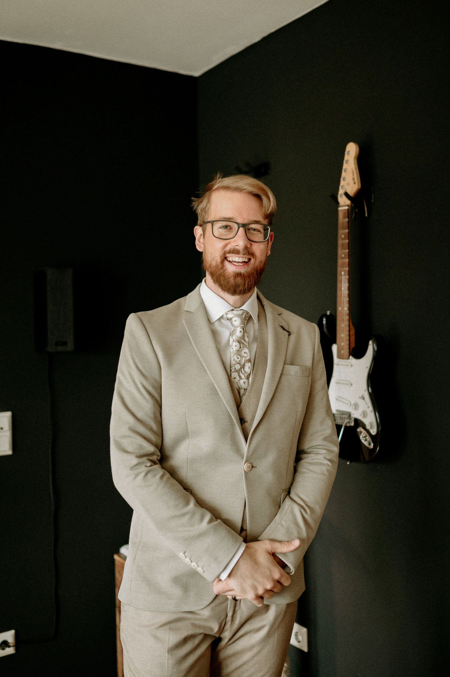 Groom in beige suit and glasses poses next to electric guitar mounted on dark wall.