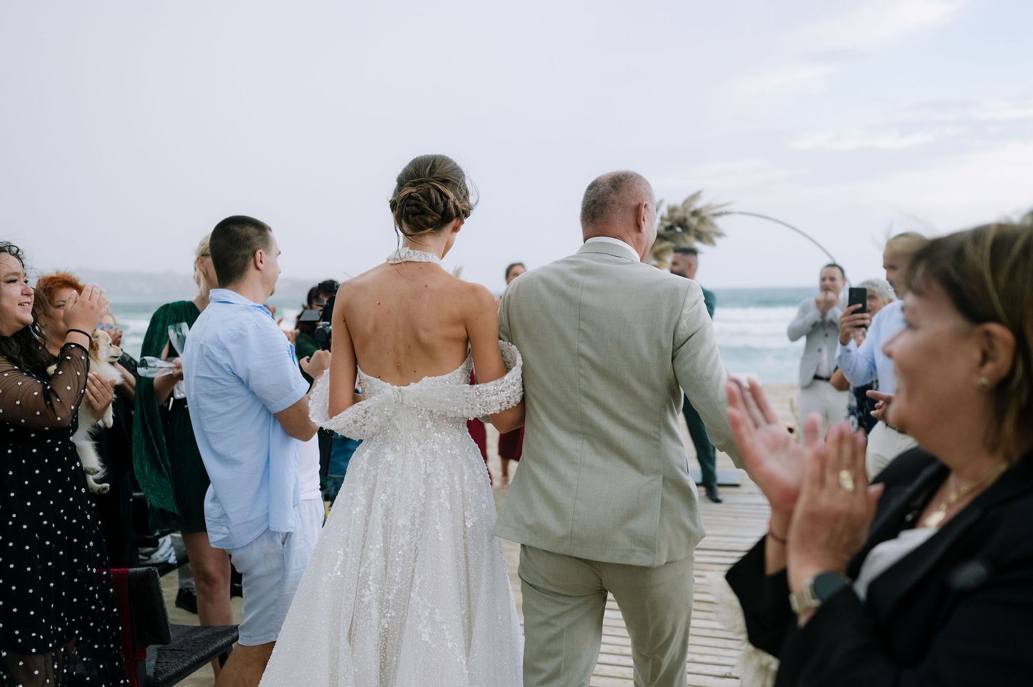Wedding guests watch as the couple walks down a beach boardwalk during the celebration.