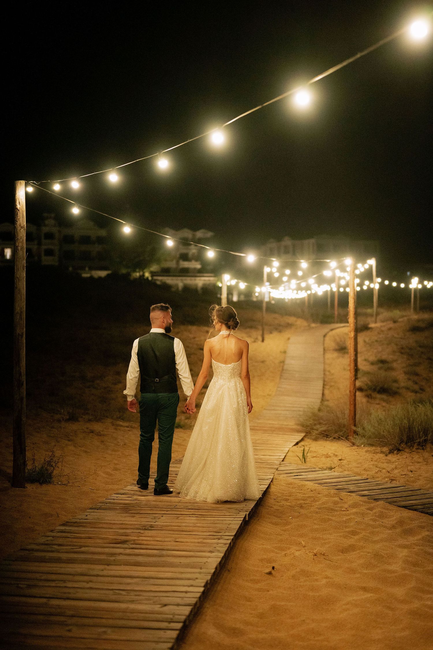 Wooden boardwalk path lit by string lights leading through desert landscape at night.
