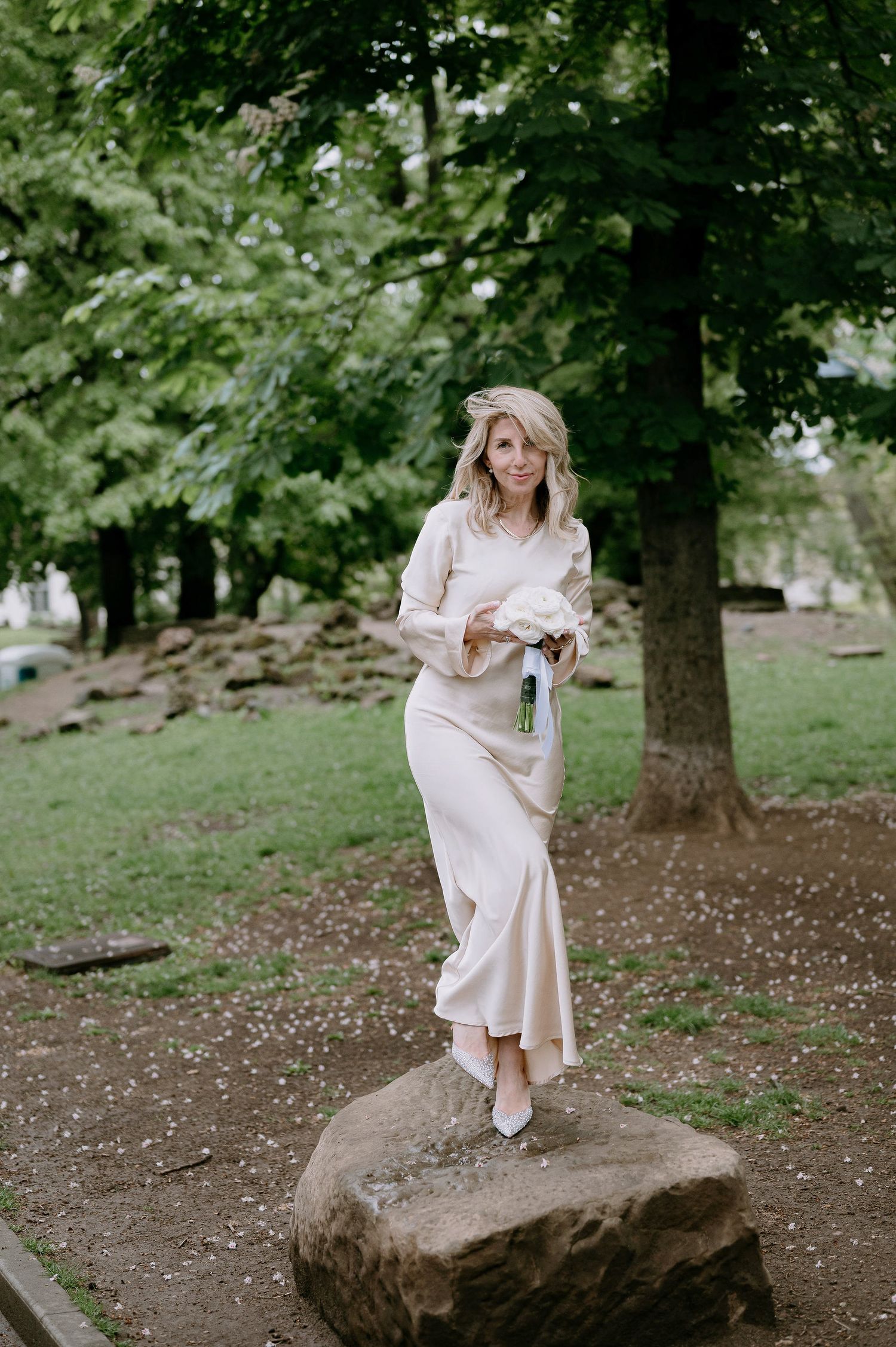 A person wearing a white dress carefully balances on a large rock in a park setting.