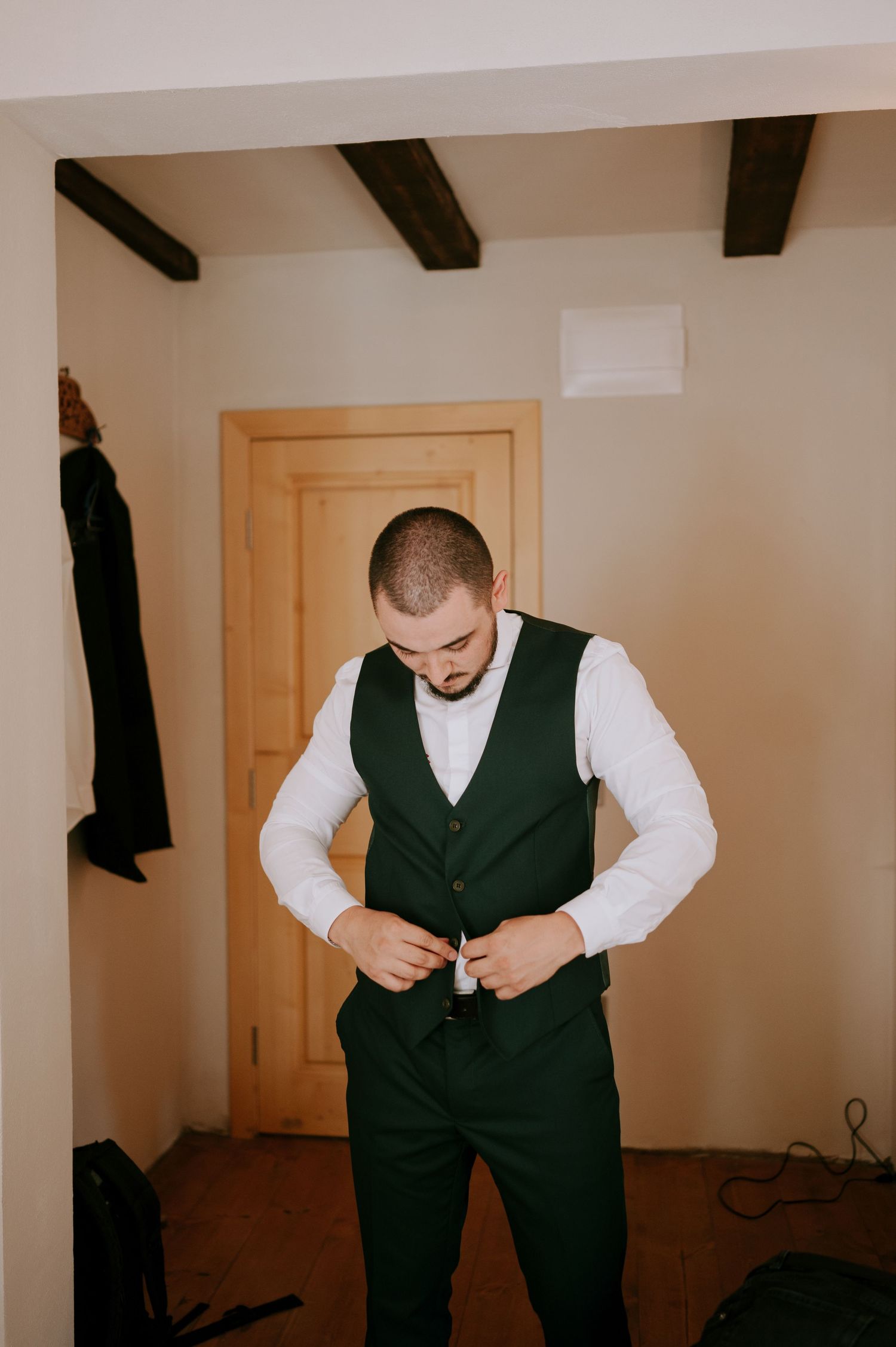 A groom adjusts his dark green vest while getting ready in a room with wooden beams.