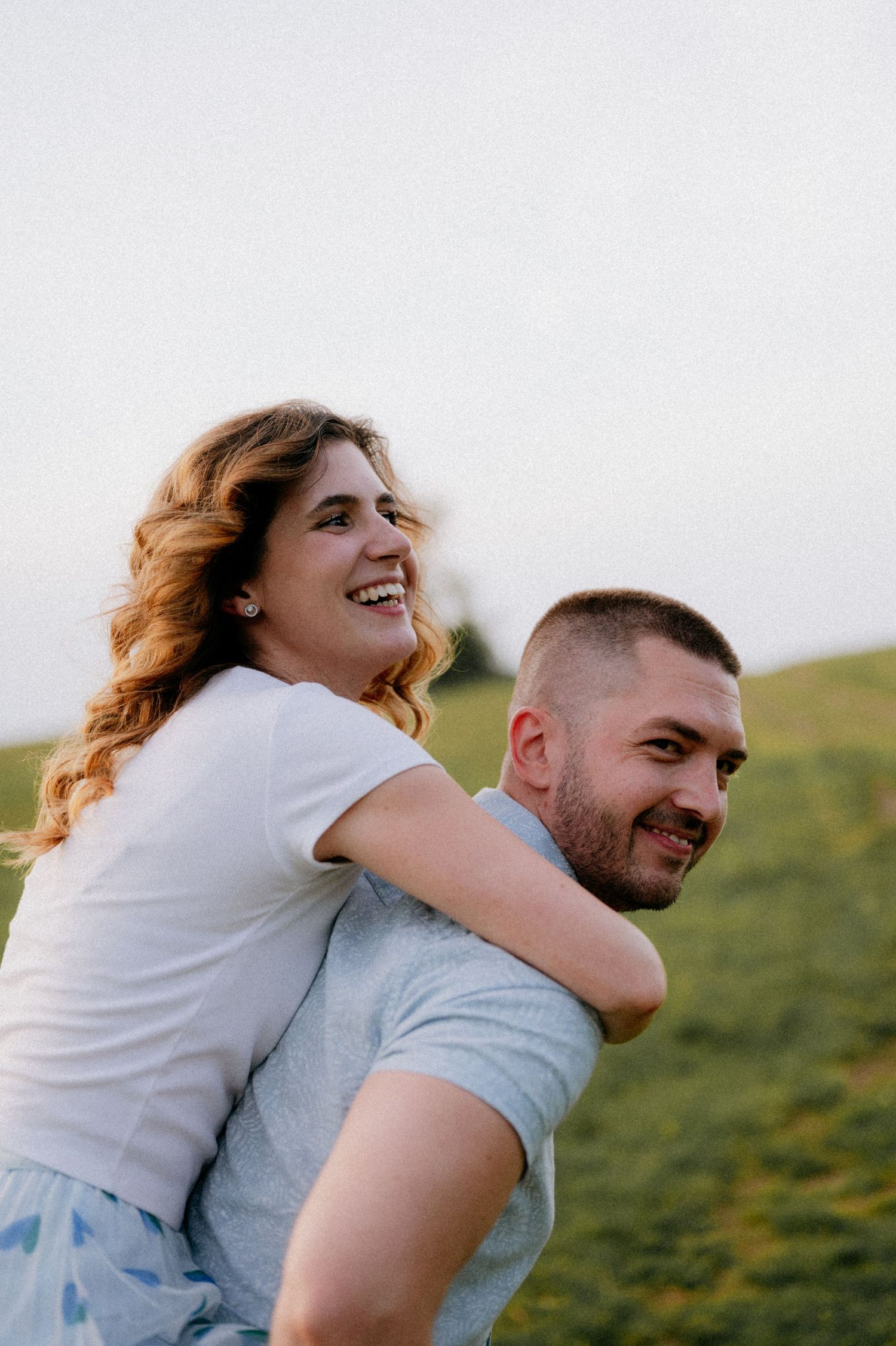 A couple embraces and laughs together on a grassy hillside during golden hour.