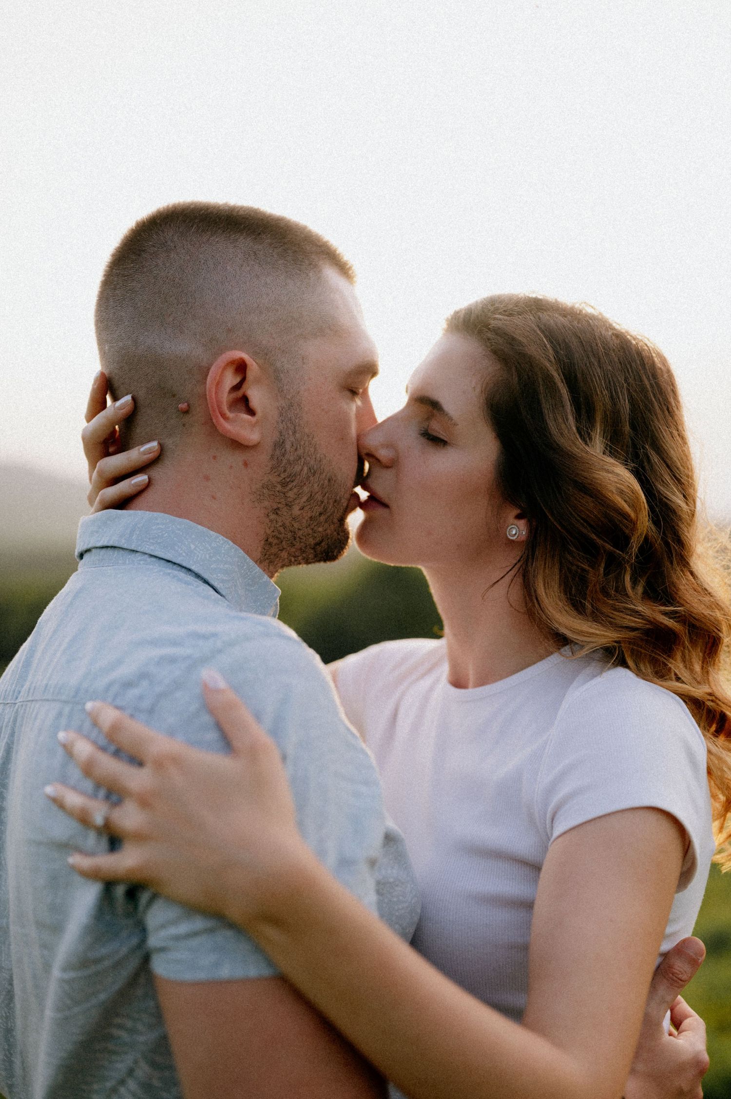 Couple embracing and kissing in casual attire during golden hour outdoors.