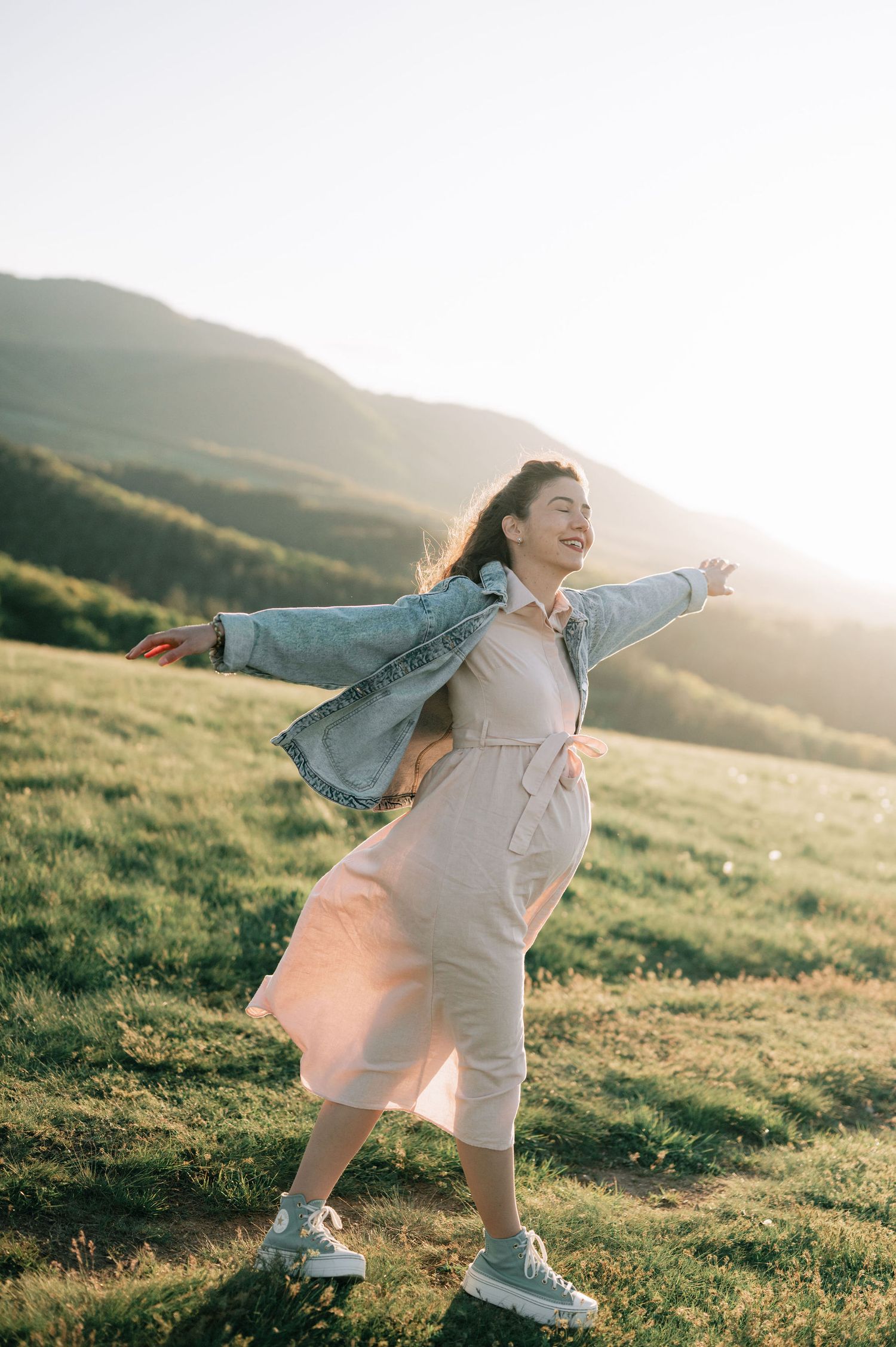 Person in denim jacket and white outfit dances joyfully in a sun-drenched field at golden hour.