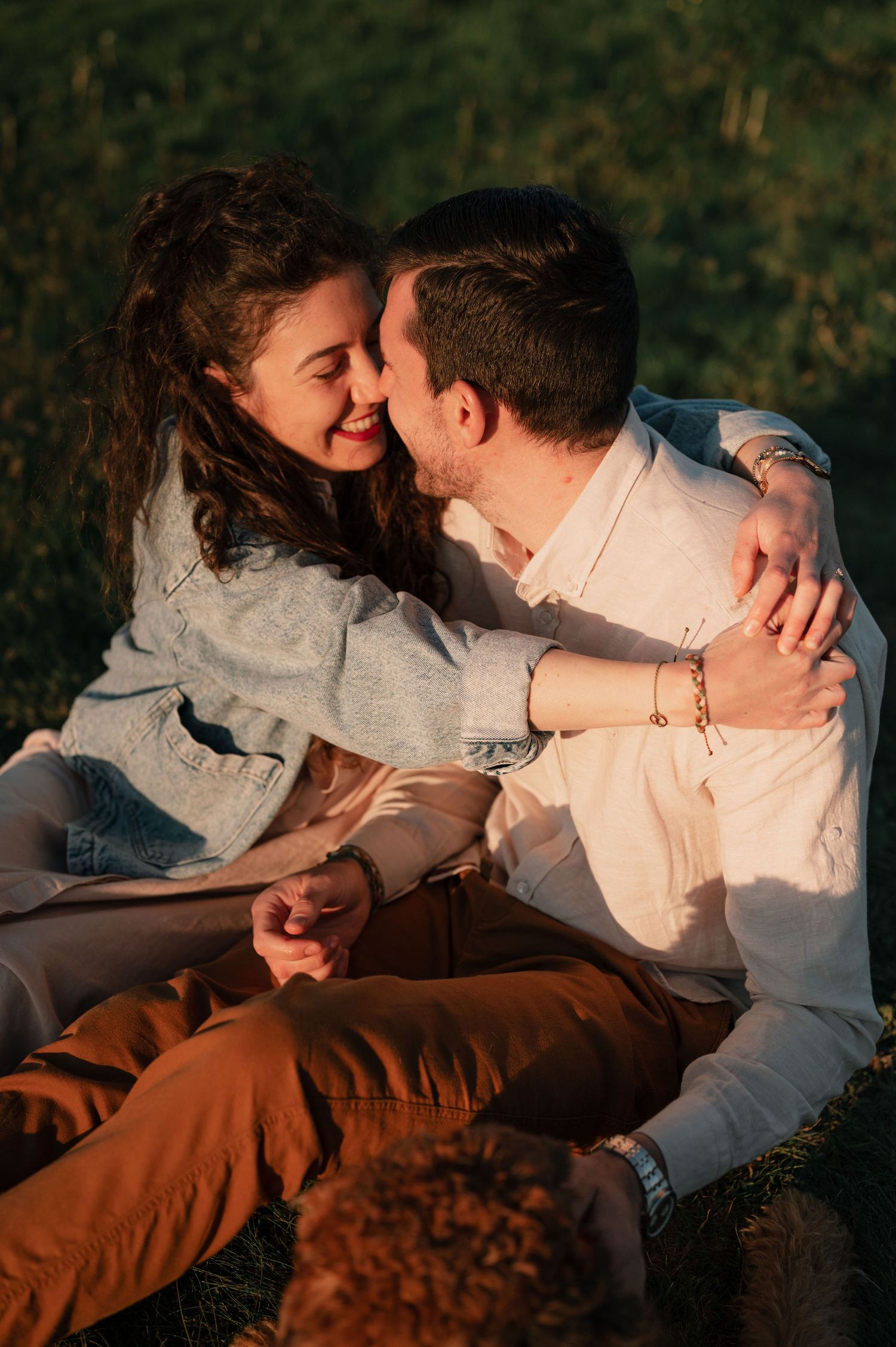 Couple shares joyful moment while sitting together on grassy hillside at sunset.