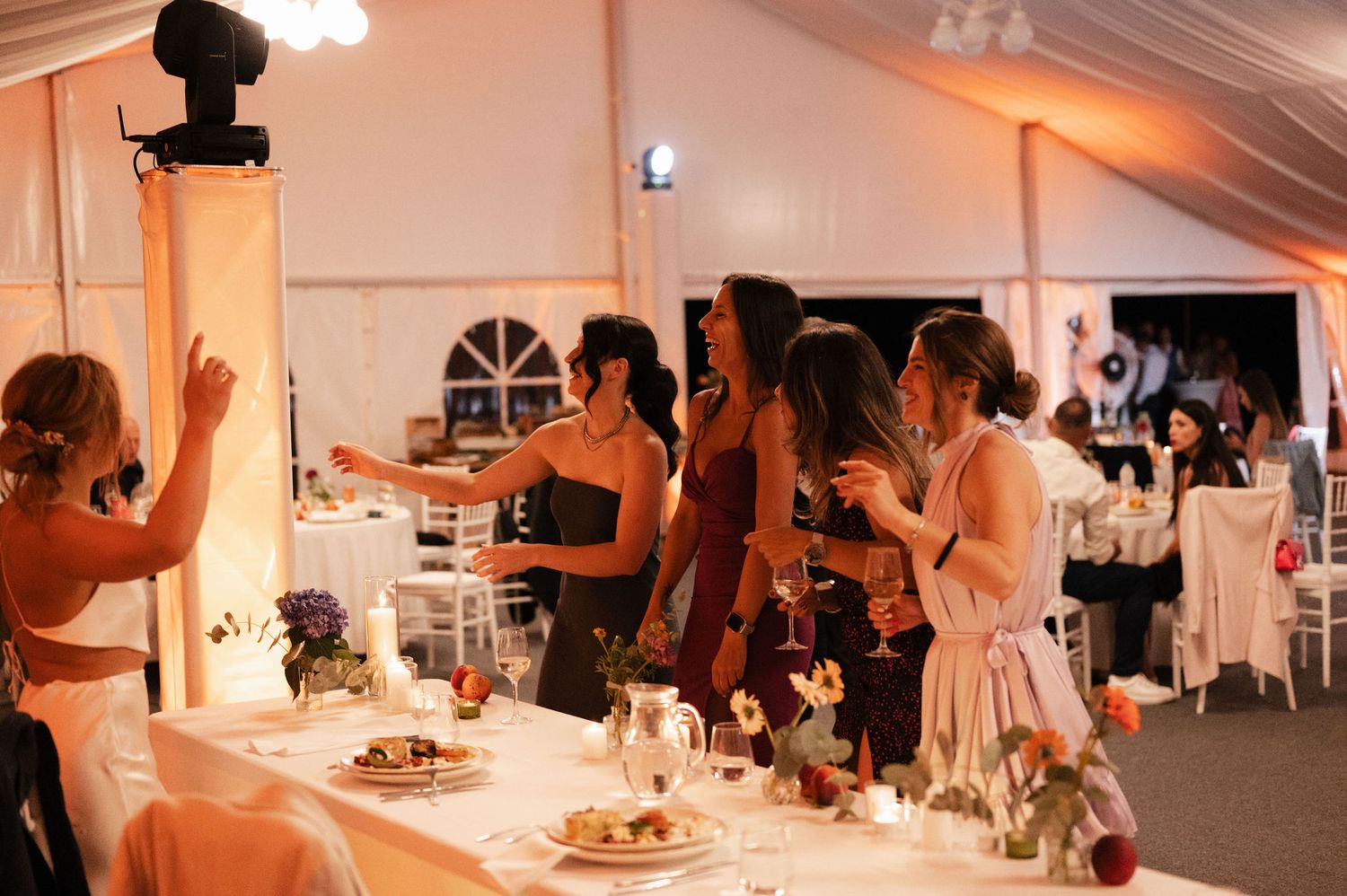 Wedding reception guests dancing and celebrating in an elegant white tent with warm lighting and decorative details.