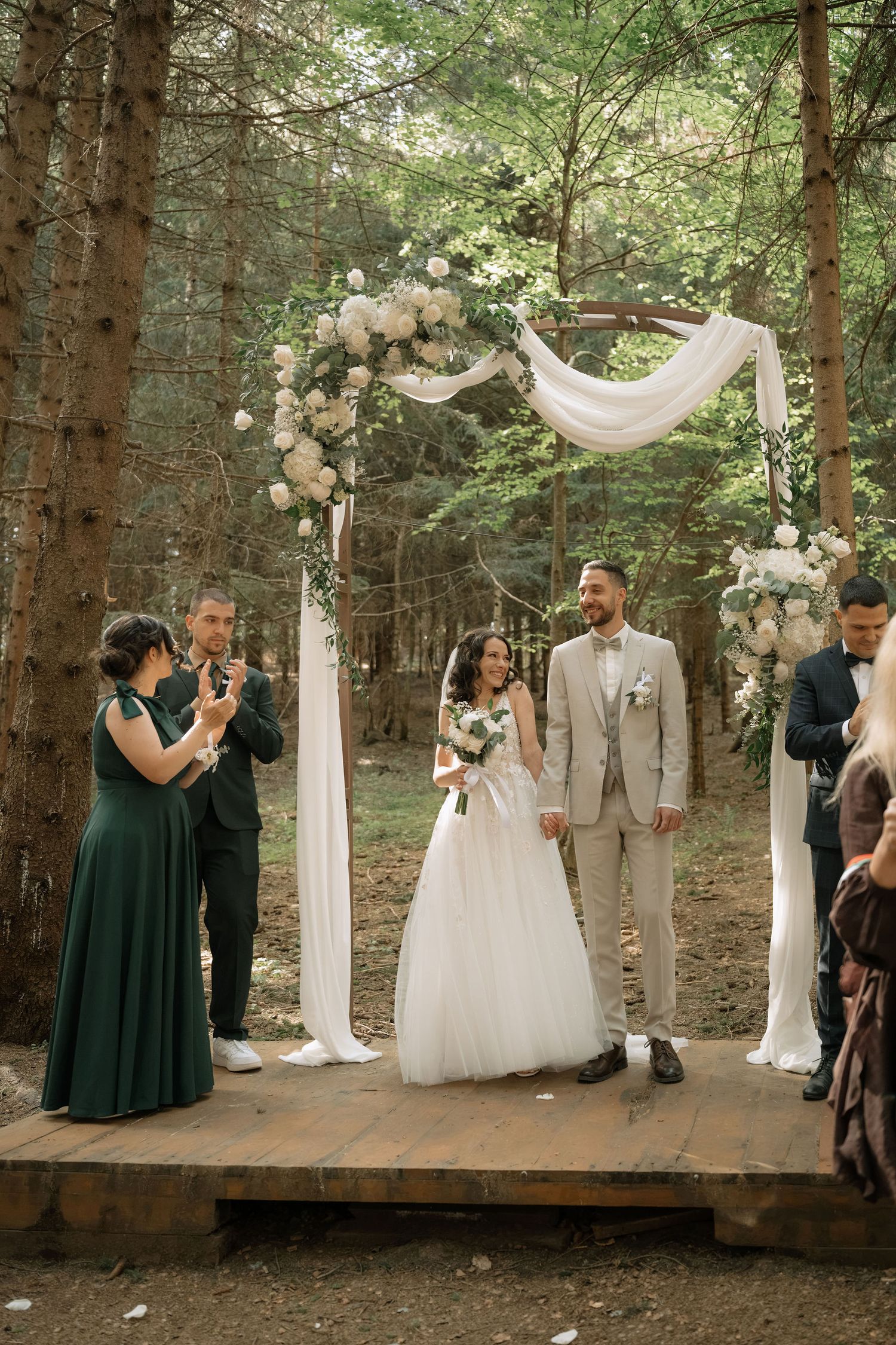 Couple exchanges vows at outdoor forest wedding ceremony under floral arch with white draping and roses.