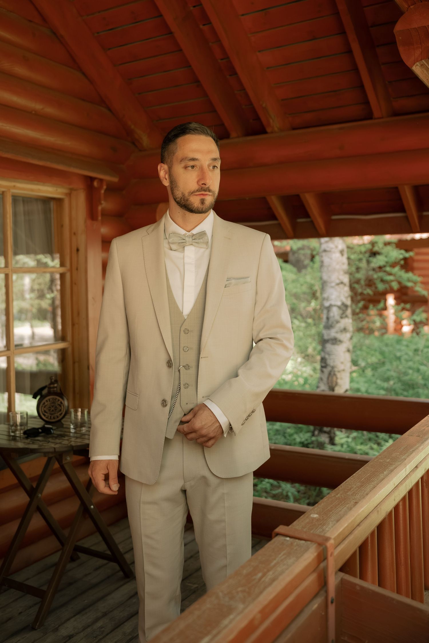 Man in beige three-piece suit with bow tie stands on wooden cabin porch surrounded by lush green trees.