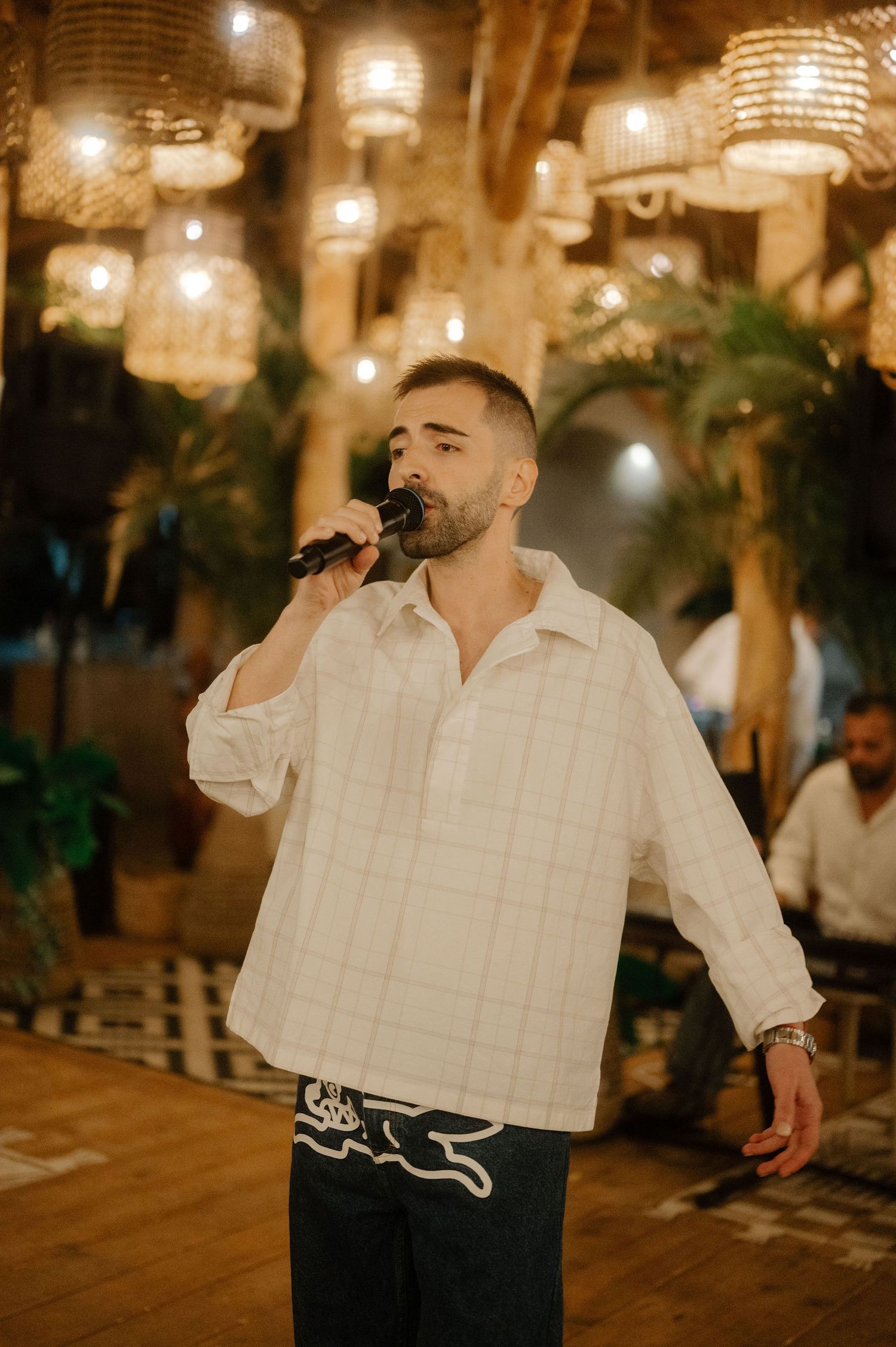 A performer sings into a microphone at an indoor venue with decorative hanging lanterns and tropical plants in the background.