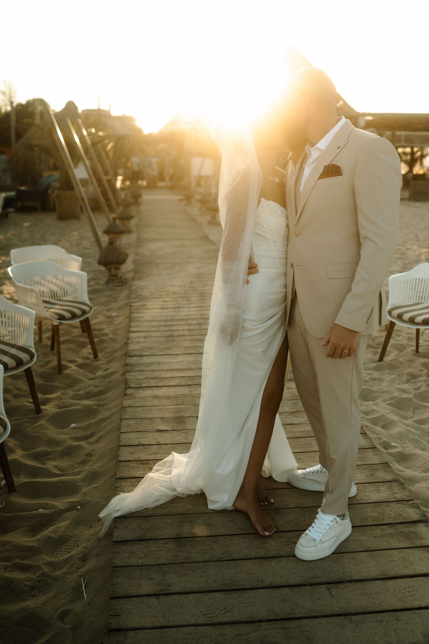 Feet of wedding couple standing on wooden boardwalk at sunset with sun flare in background.