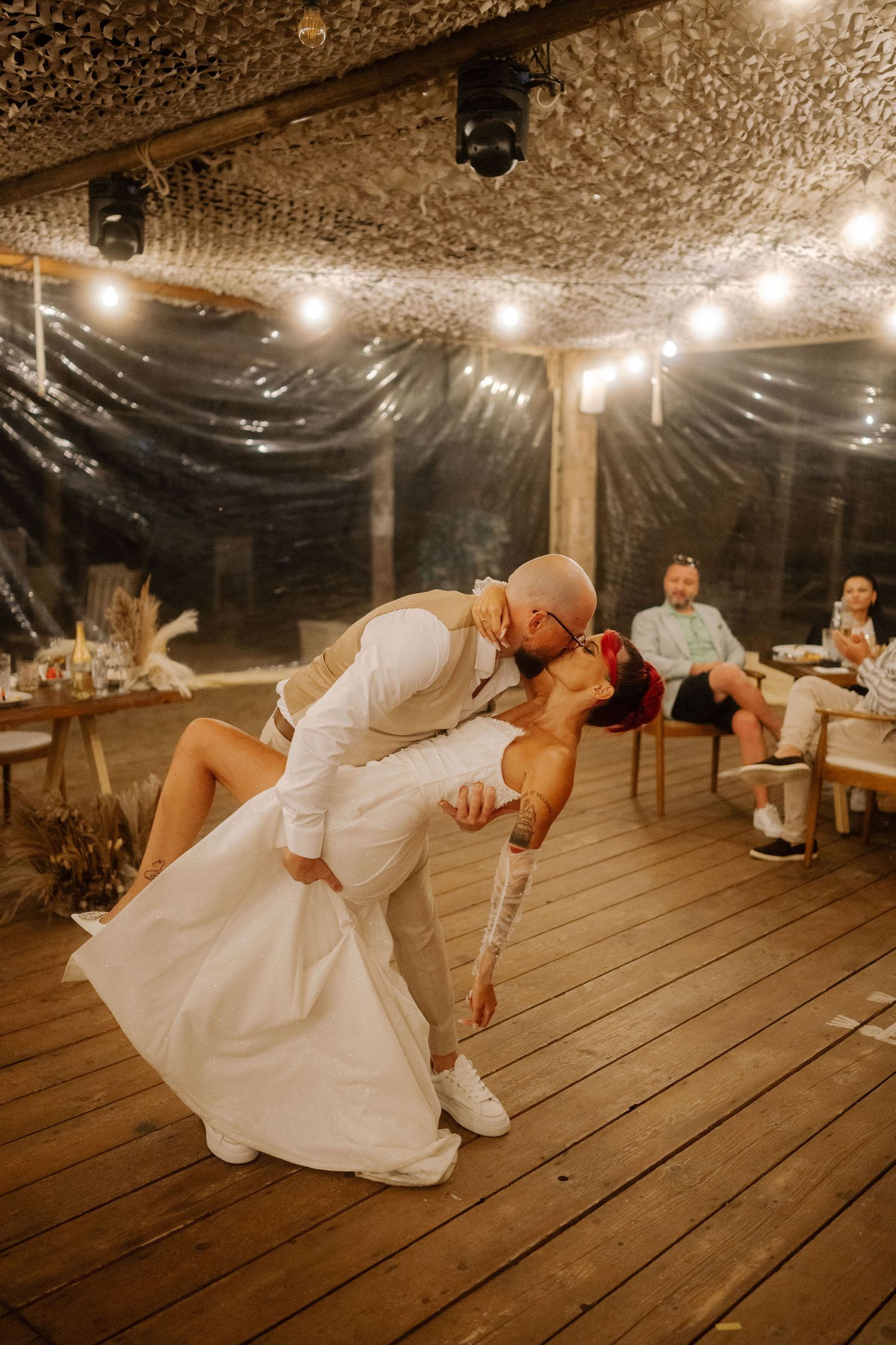 A couple performs a romantic dip dance move on a rustic wooden deck under string lights at an evening celebration.