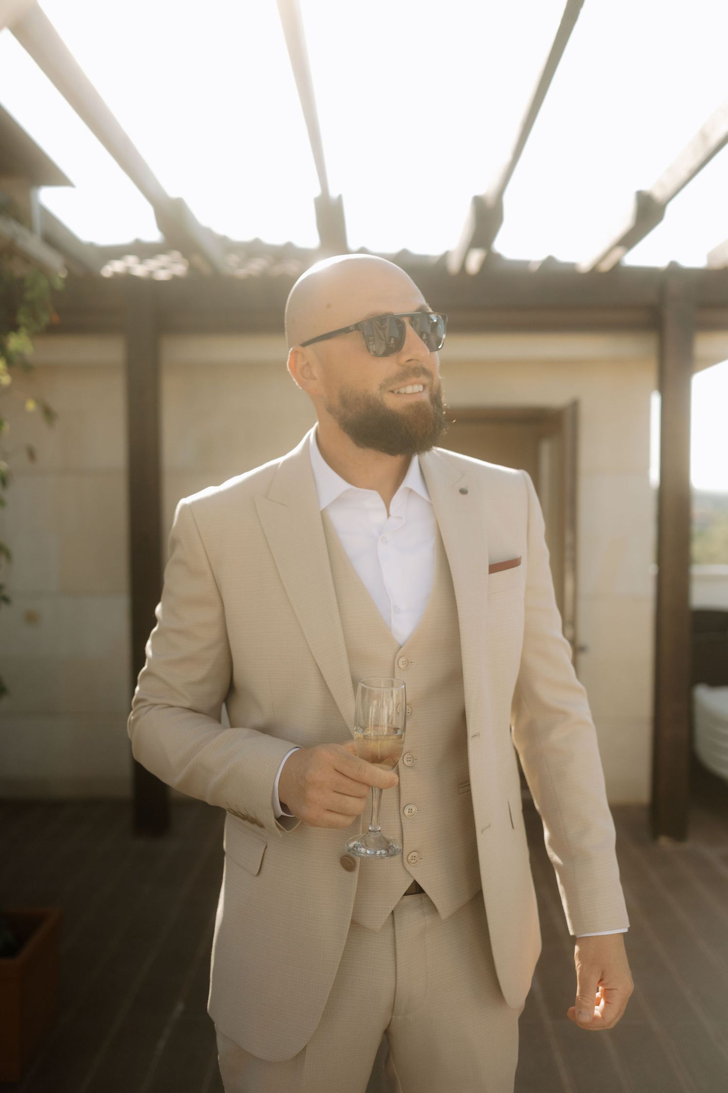 A person in a beige three-piece suit and sunglasses holds a champagne glass during a sunset photoshoot.