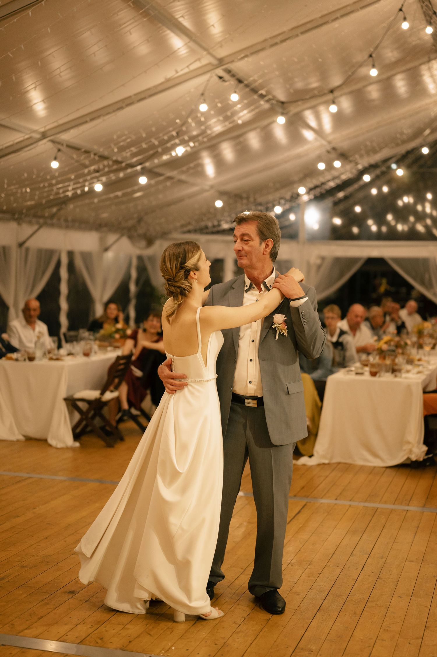 A couple shares a romantic dance together on a wooden floor under a tent with string lights at an evening reception.