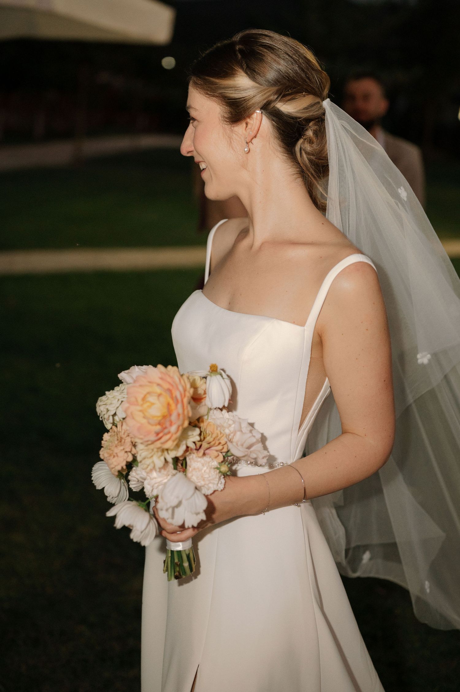 A bride in a simple white dress holds a soft peach and white floral bouquet against a dark background.