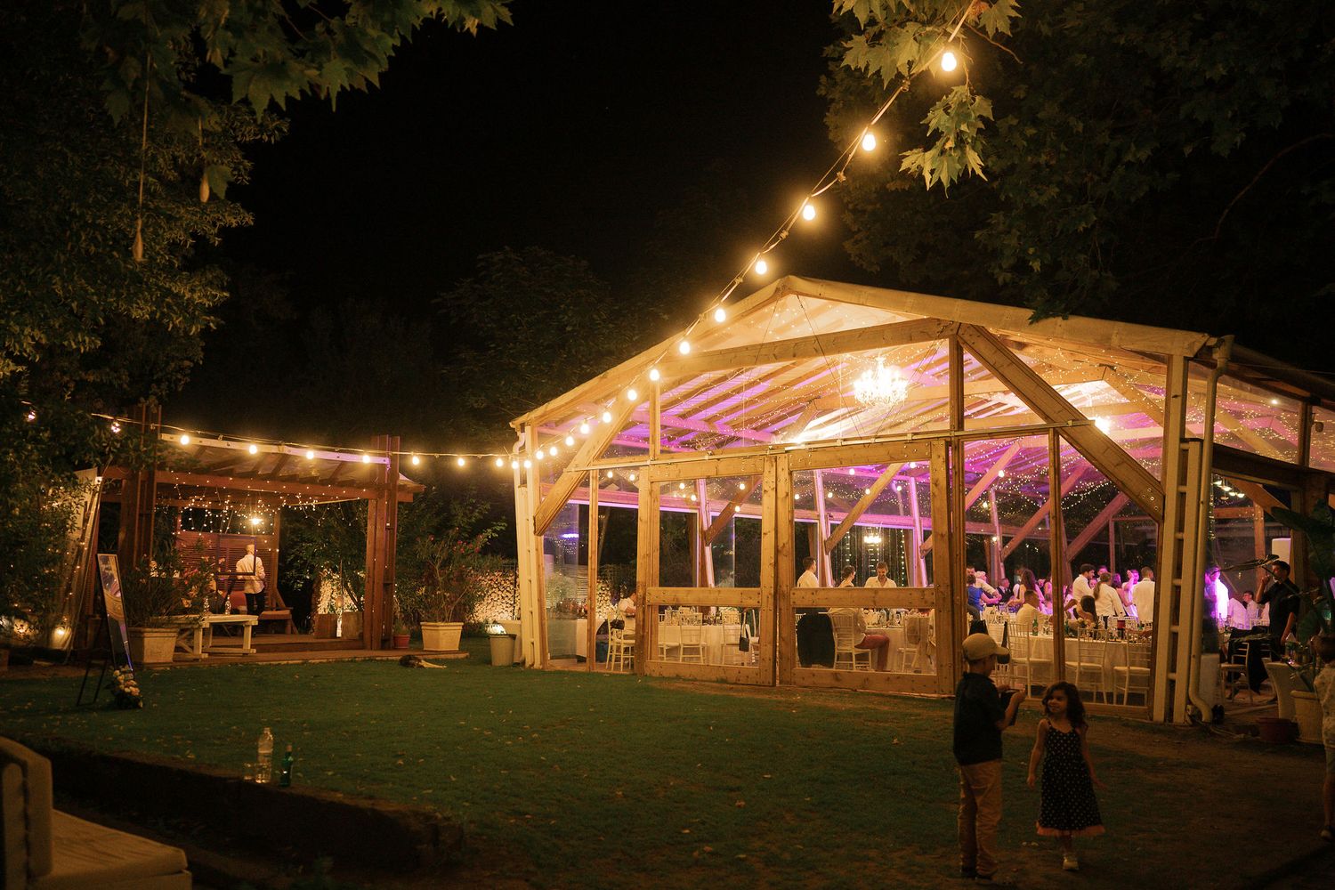 Illuminated wedding tent with string lights glowing warmly at night in an outdoor garden setting.