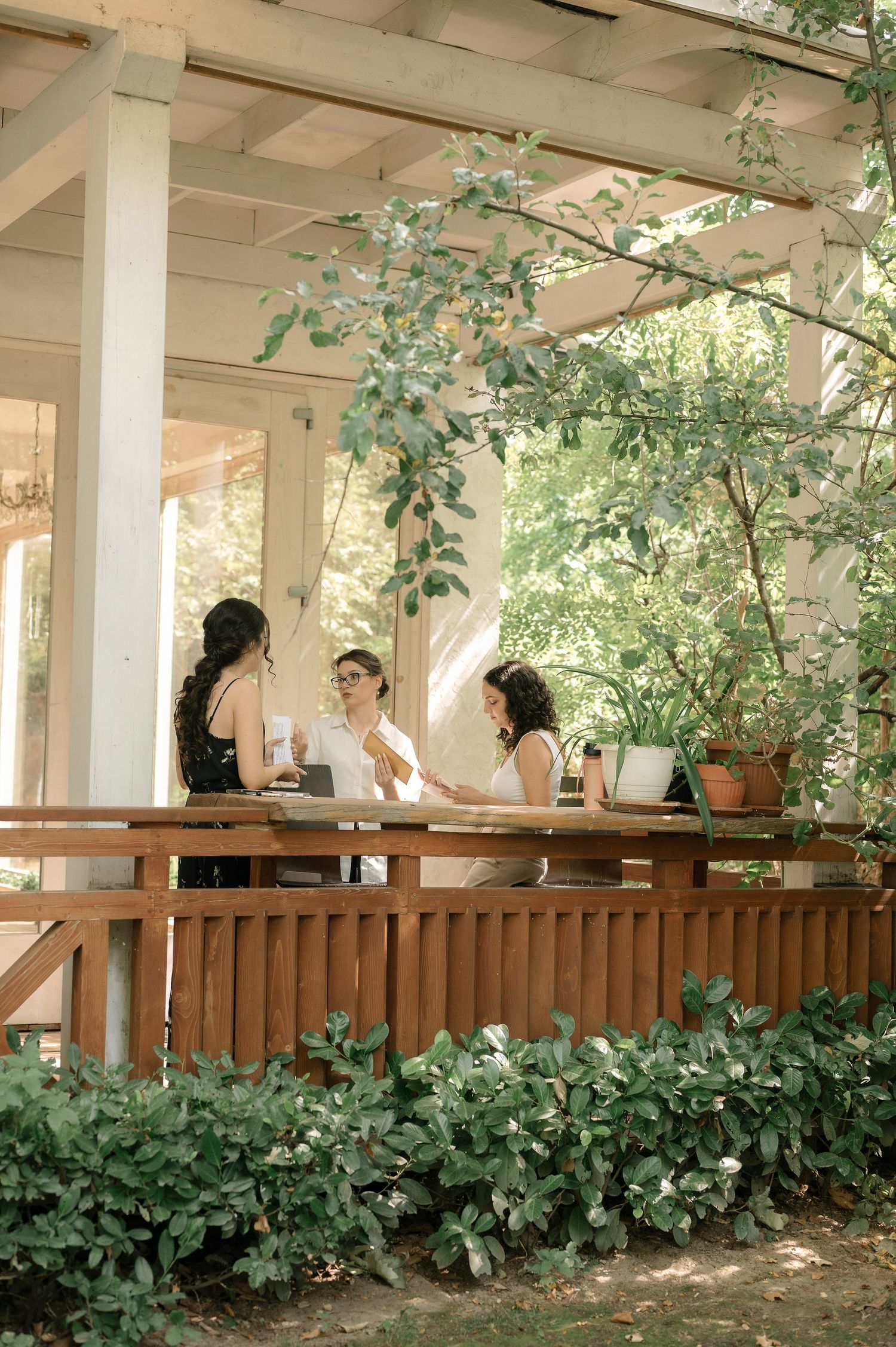 Three people enjoying drinks and conversation on a rustic wooden porch surrounded by lush greenery on a sunny afternoon.