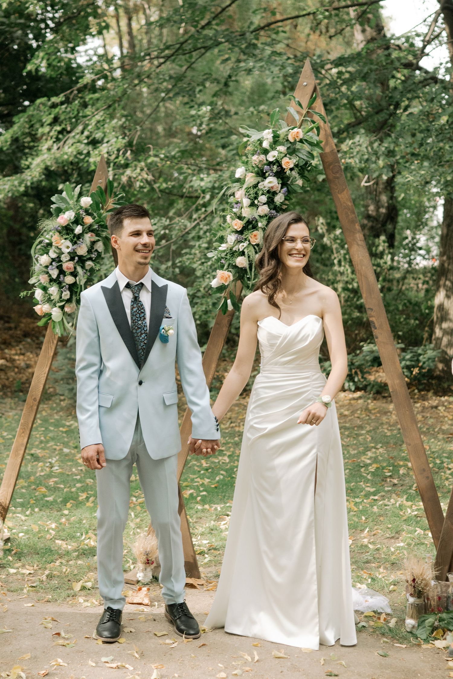 A couple in wedding attire walks through a wooded outdoor setting with scattered flower petals on the ground.