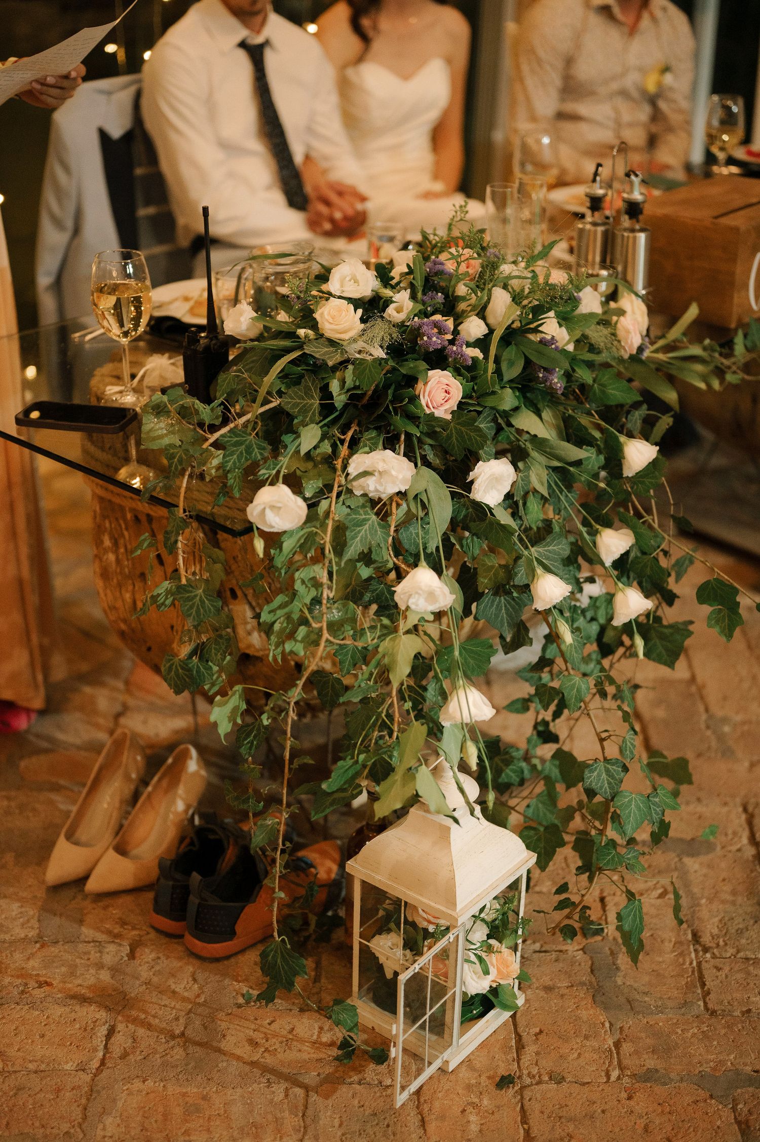 Elegant wedding reception decor with cascading white roses and greenery arrangement next to a white lantern on stone floor.