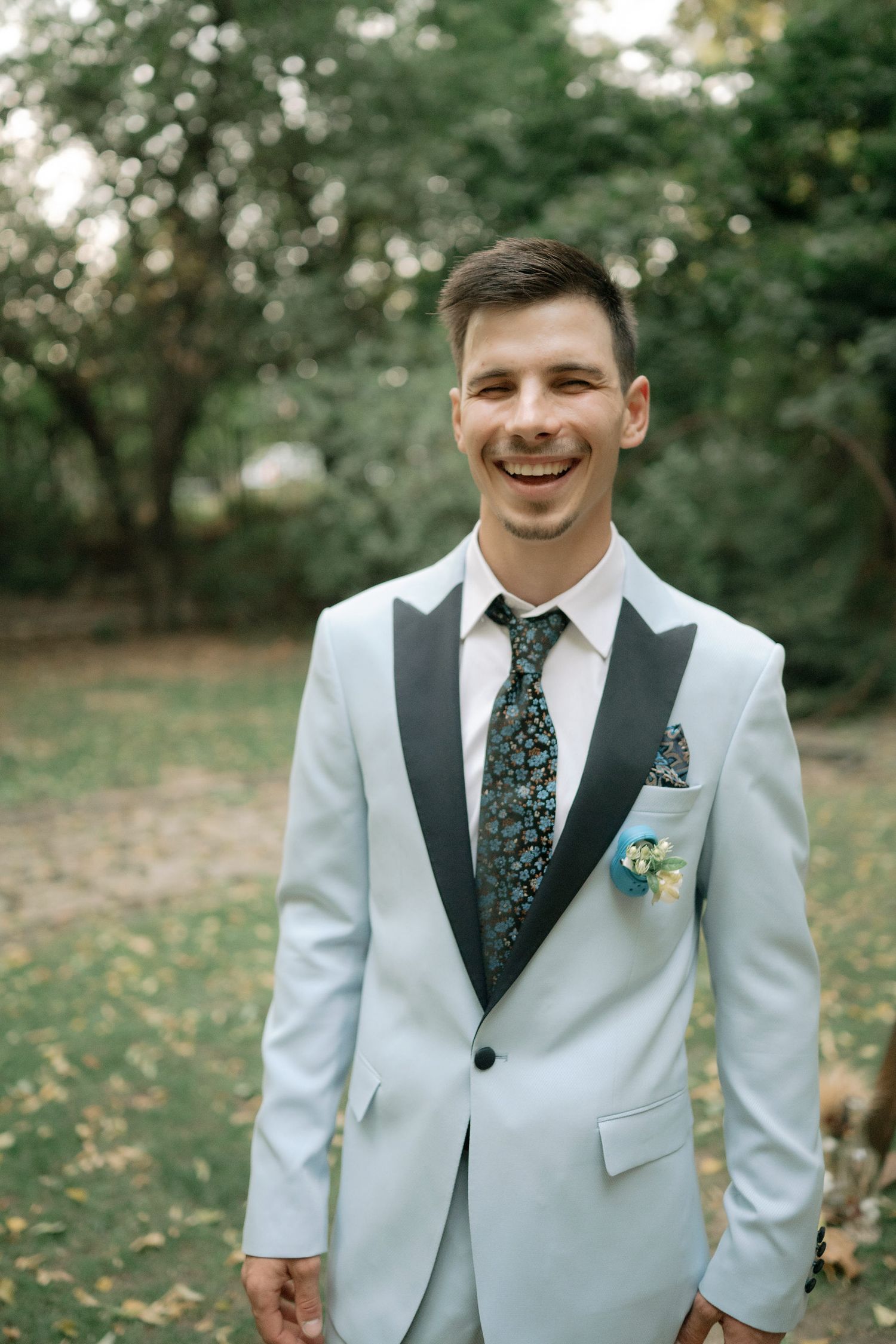 A person wearing a light gray tuxedo with black lapels and floral tie smiles outdoors in a garden setting.
