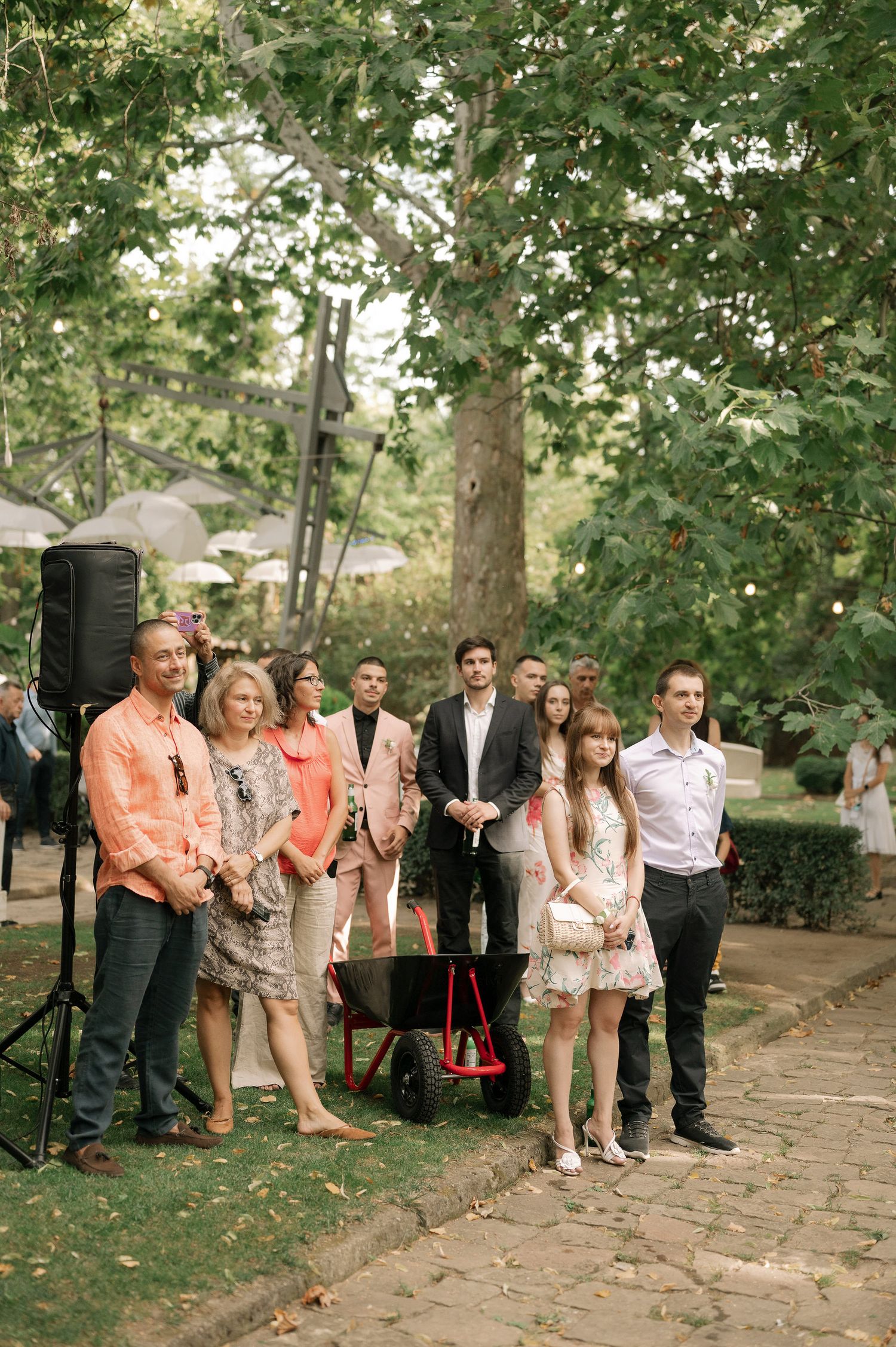 Wedding party guests line up along a stone pathway in a garden setting wearing coral and neutral colored attire.