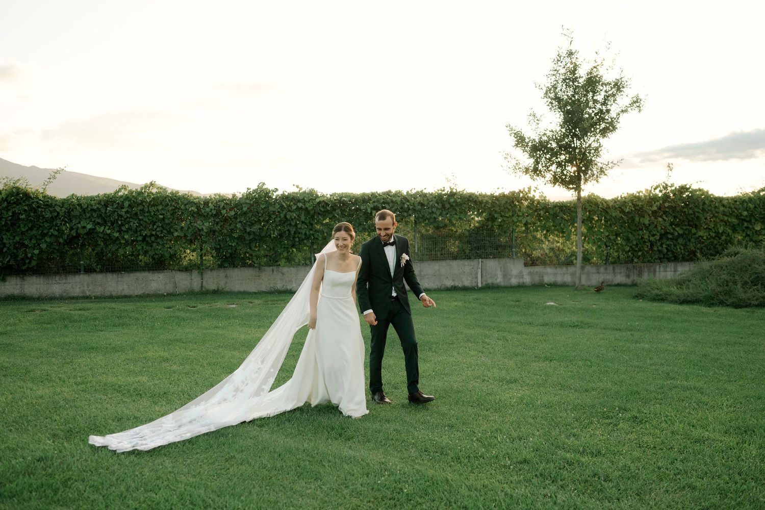 A couple in wedding attire stands together on a lush green lawn during a sunset ceremony.