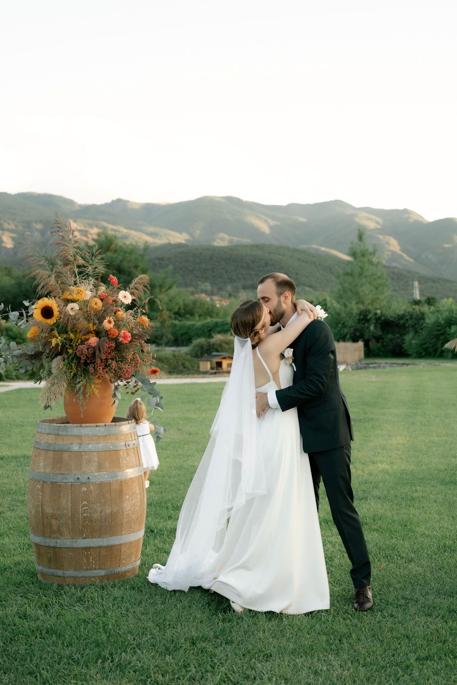 A romantic outdoor wedding scene with a rustic wine barrel and sunflower decor in a mountain meadow setting.