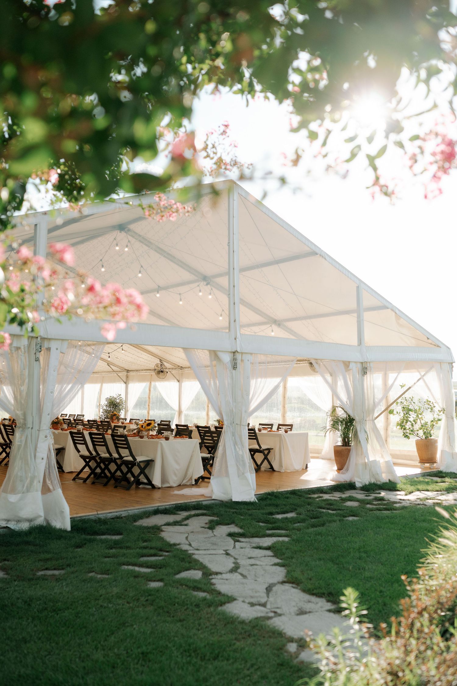 White wedding tent pavilion set up on a lush green lawn with flowering trees and shaded outdoor seating areas.