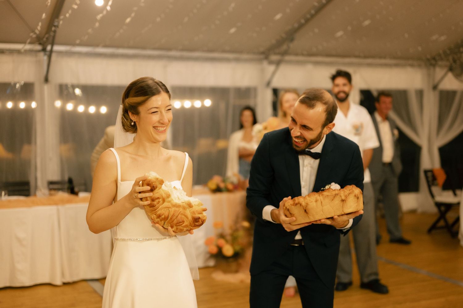 Wedding guests dance together in a rustic reception venue decorated with string lights and white drapes.